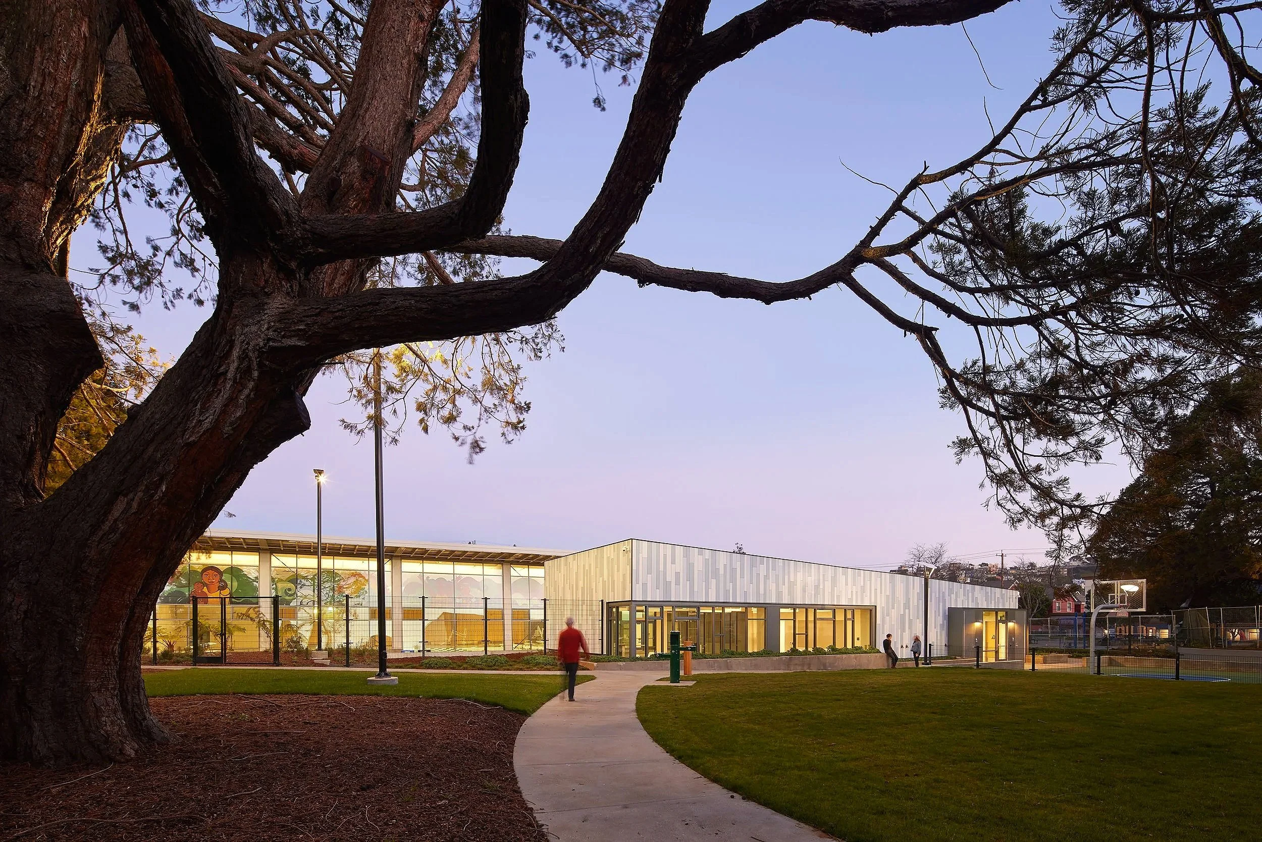 Modern school building with large windows and a curved pathway, set in a park-like area with trees and a basketball court.