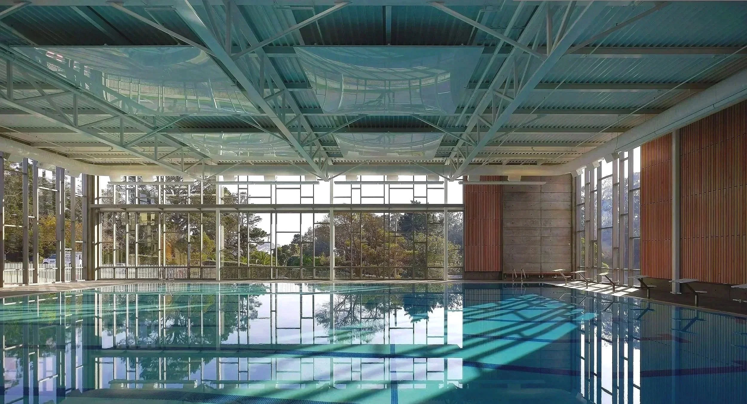 Indoor swimming pool with large glass windows and ceiling, natural sunlight, and trees outside, modern design with wood and concrete walls.