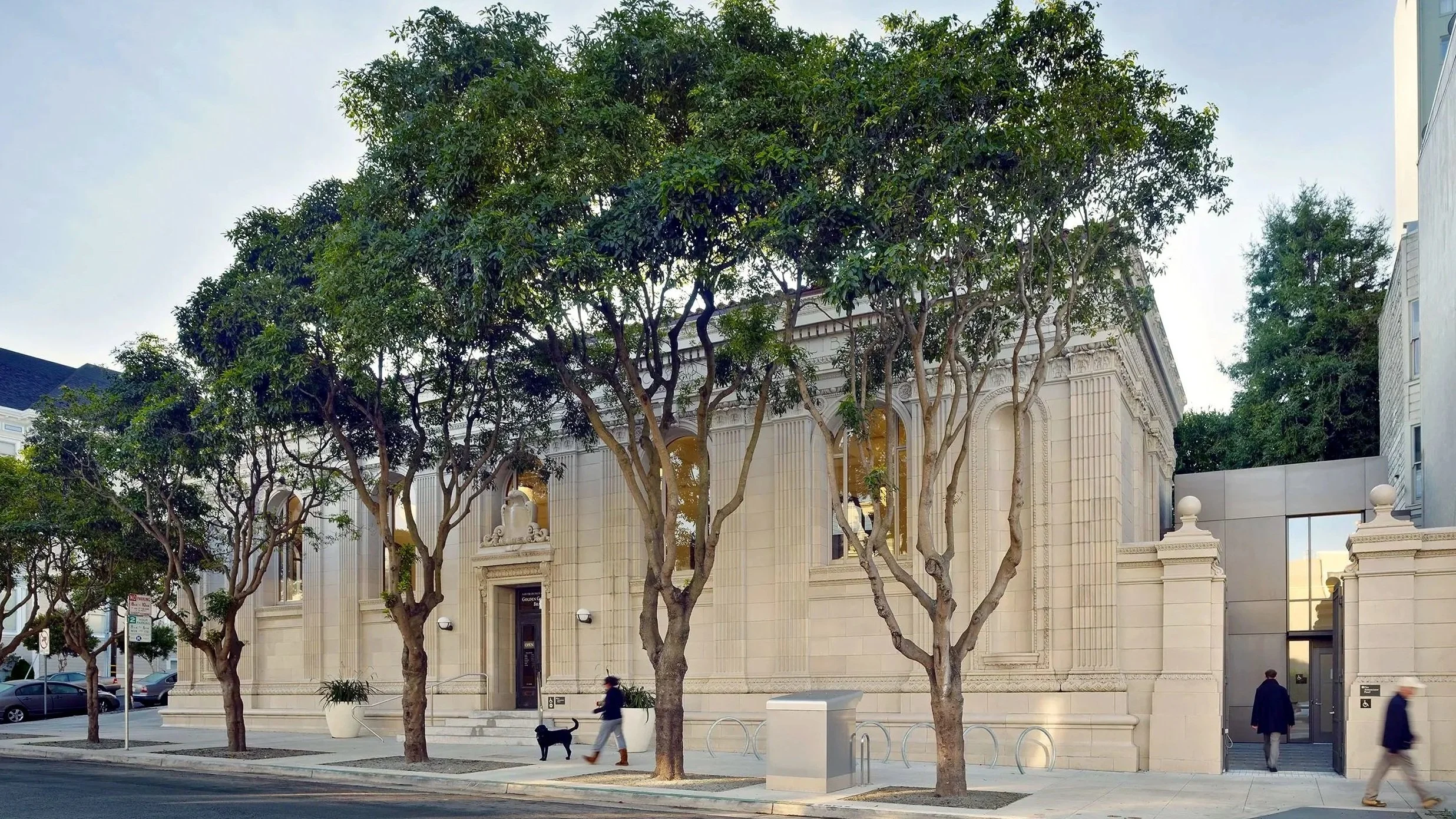 A city street scene featuring a large, ornate stone building with tall, arched windows, trees lining the sidewalk, and pedestrians walking by.