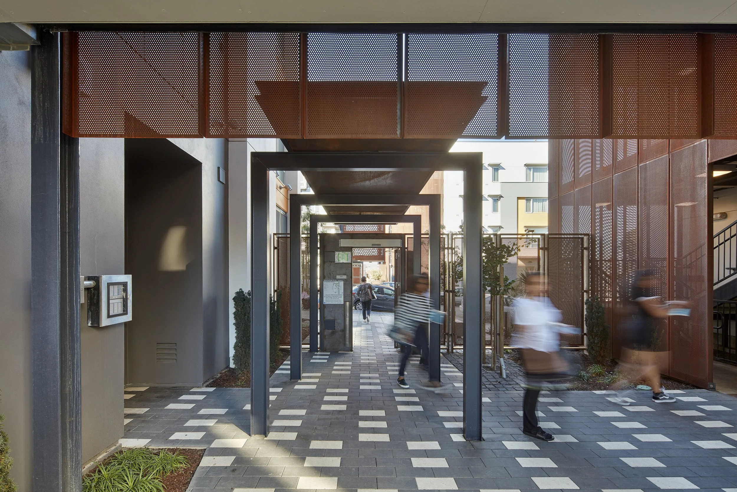 People walking through a modern outdoor corridor with metal archways and textured brick flooring front of a residential building.