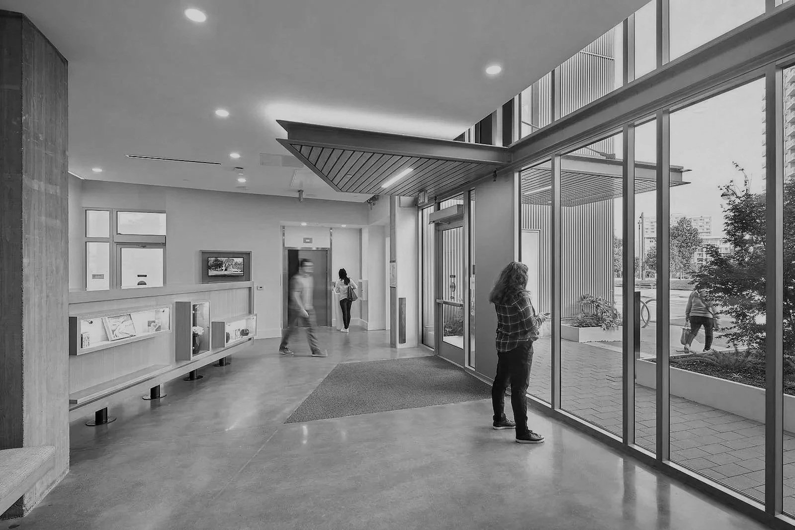 People inside a modern building lobby with large glass windows and an elevator, watching outside pedestrians.
