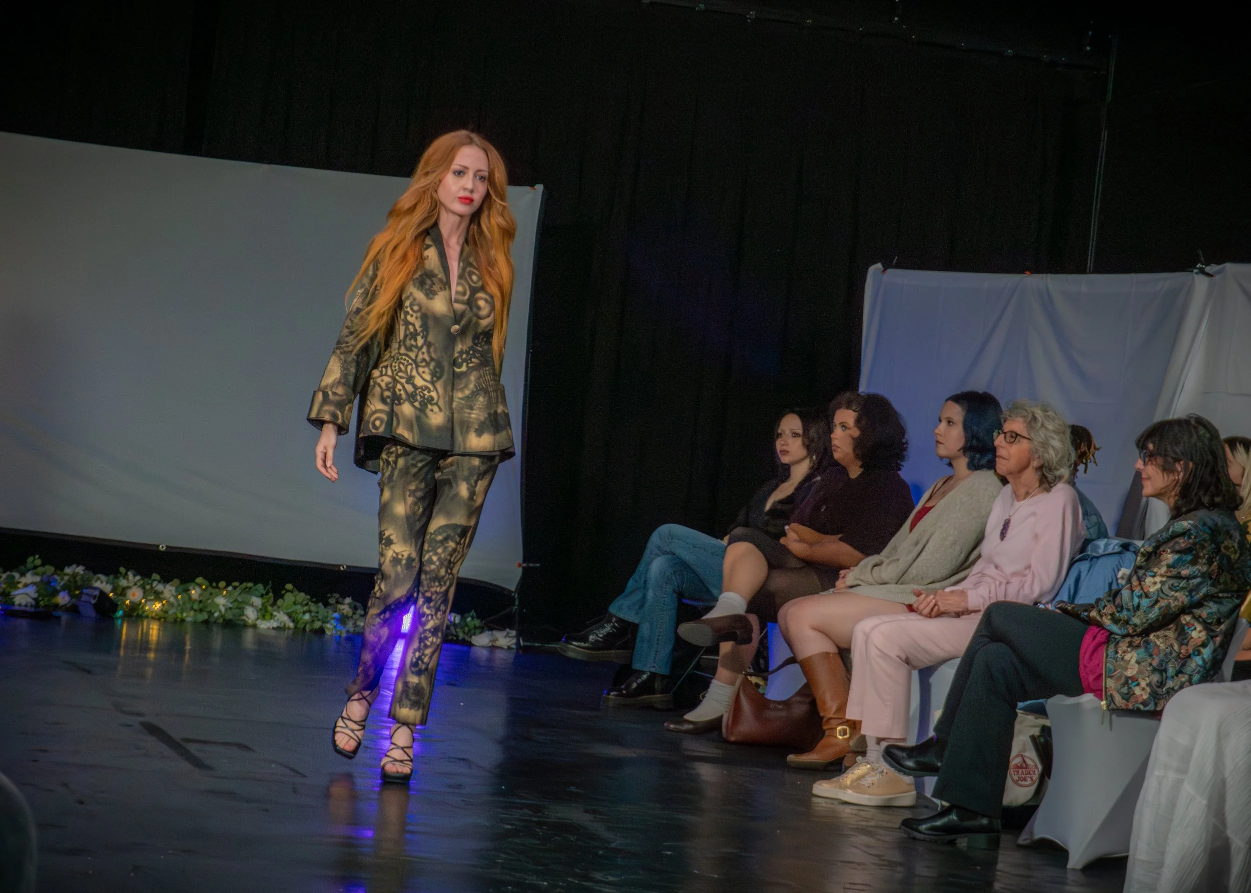 A woman with long red hair walking on a fashion runway wearing a matching outfit with a black and gold pattern. Several seated women watch the show, with some wearing glasses and fashionable clothing, and a black curtain and white presentation panels