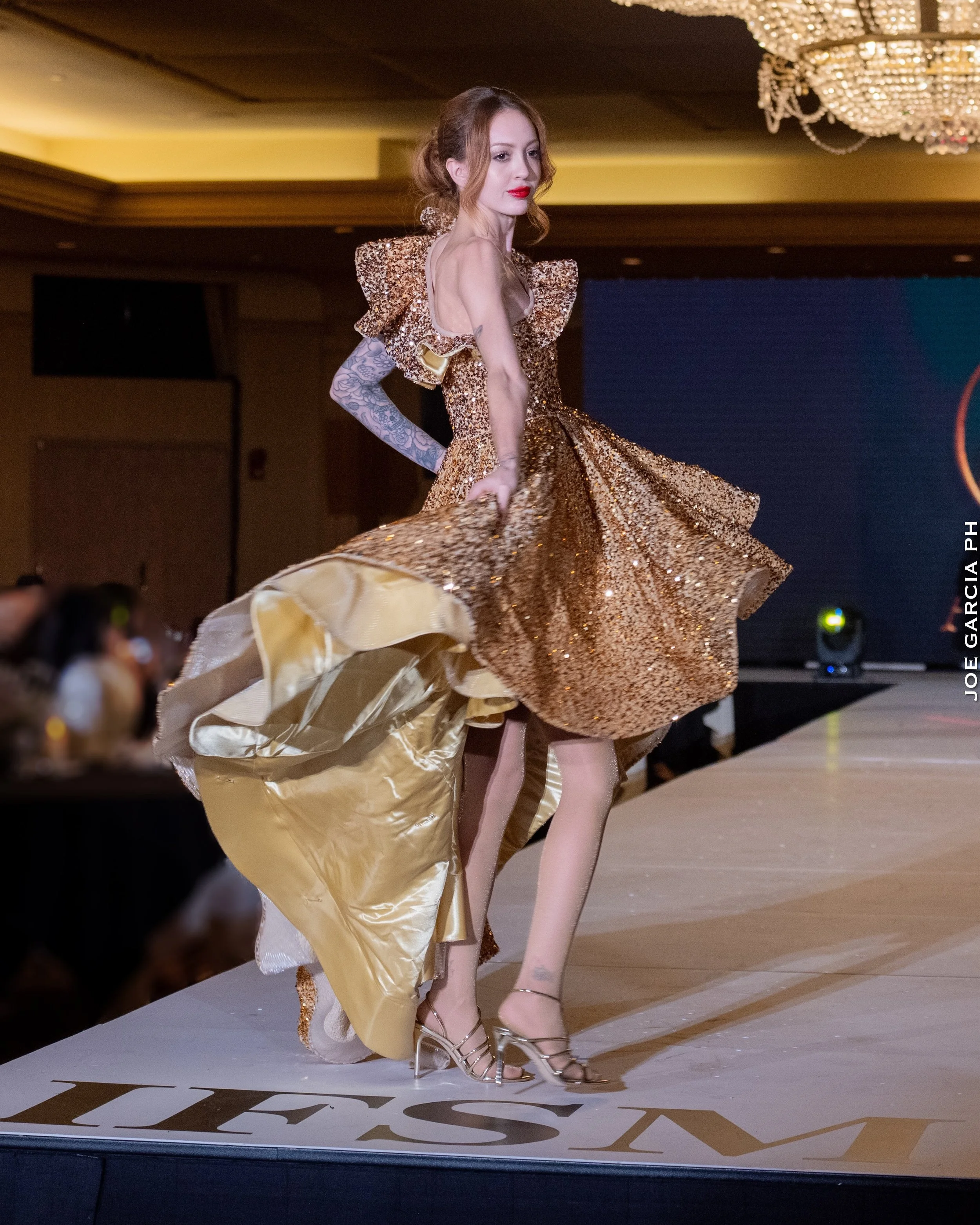 A fashion model walking on a runway in a gold, glittery gown with ruffled shoulders and a flowing, shiny skirt.