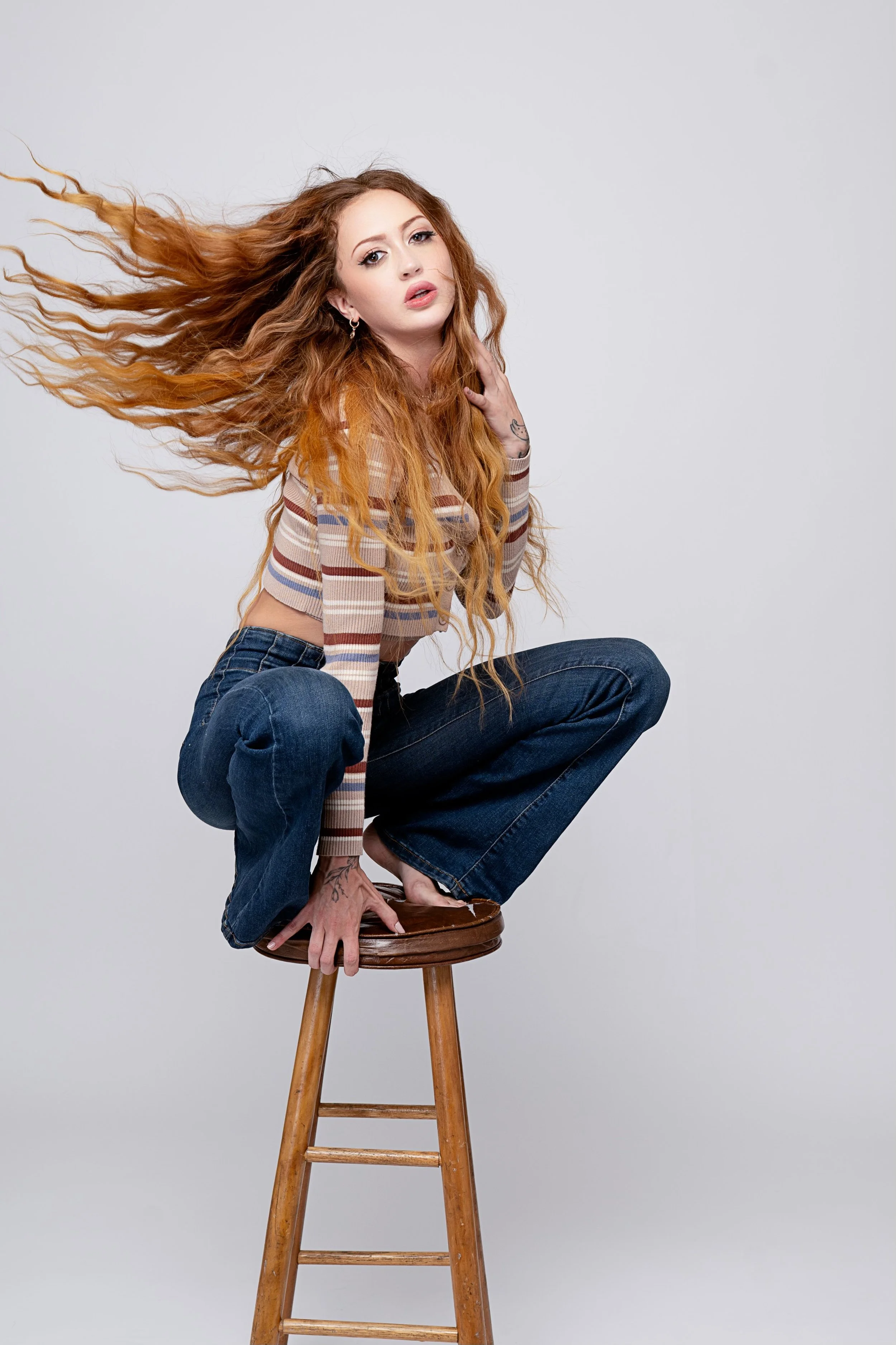 A young woman with long, wavy red hair squatting on a wooden stool against a plain white background.