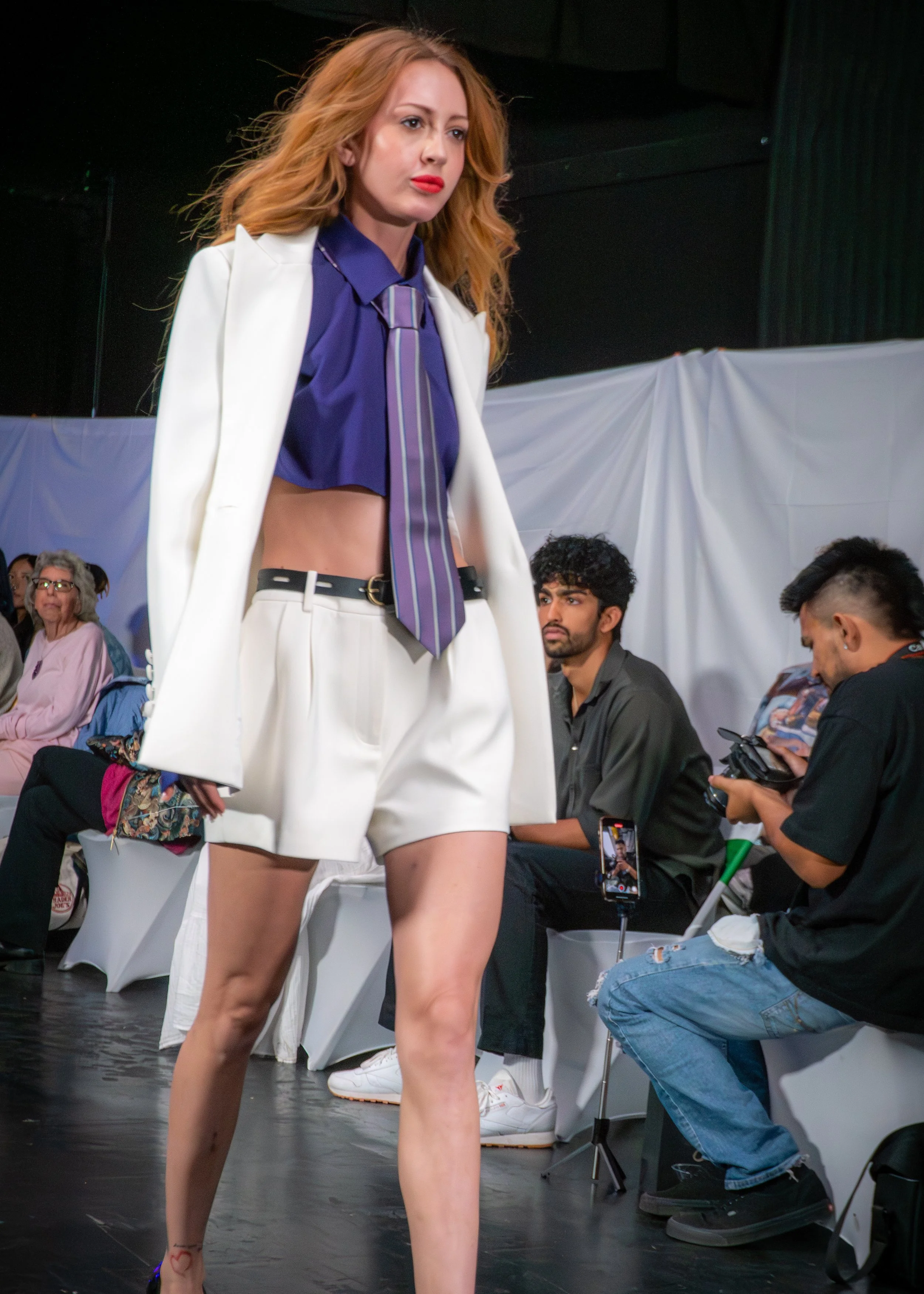 A female model walking on a runway wearing a white blazer, purple shirt, striped tie, and white shorts, with attendees seated on either side watching the show.