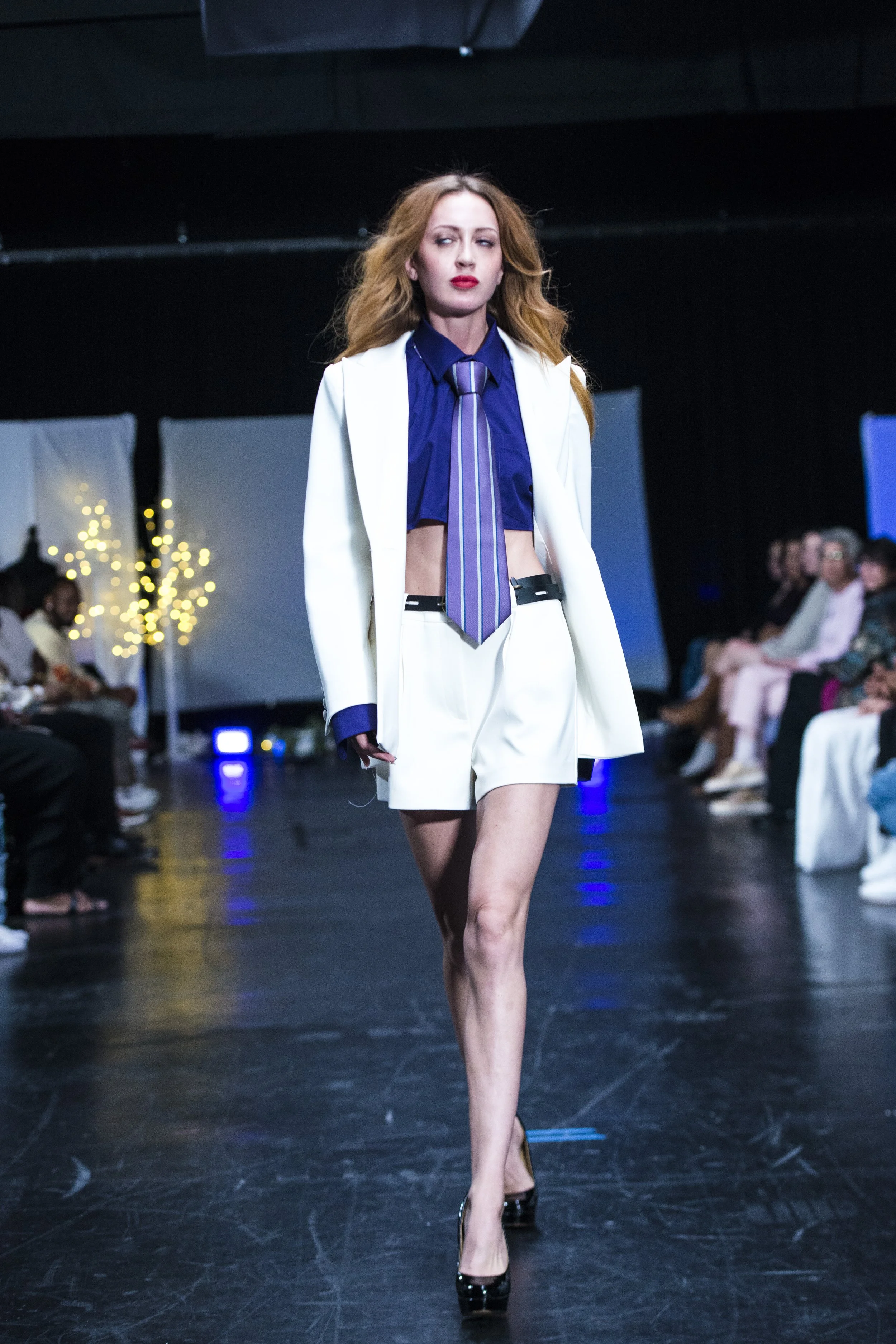 A female model walking on a runway at a fashion show, wearing a white blazer, matching shorts, a blue shirt, and a striped tie, with audience members seated on both sides of the runway.