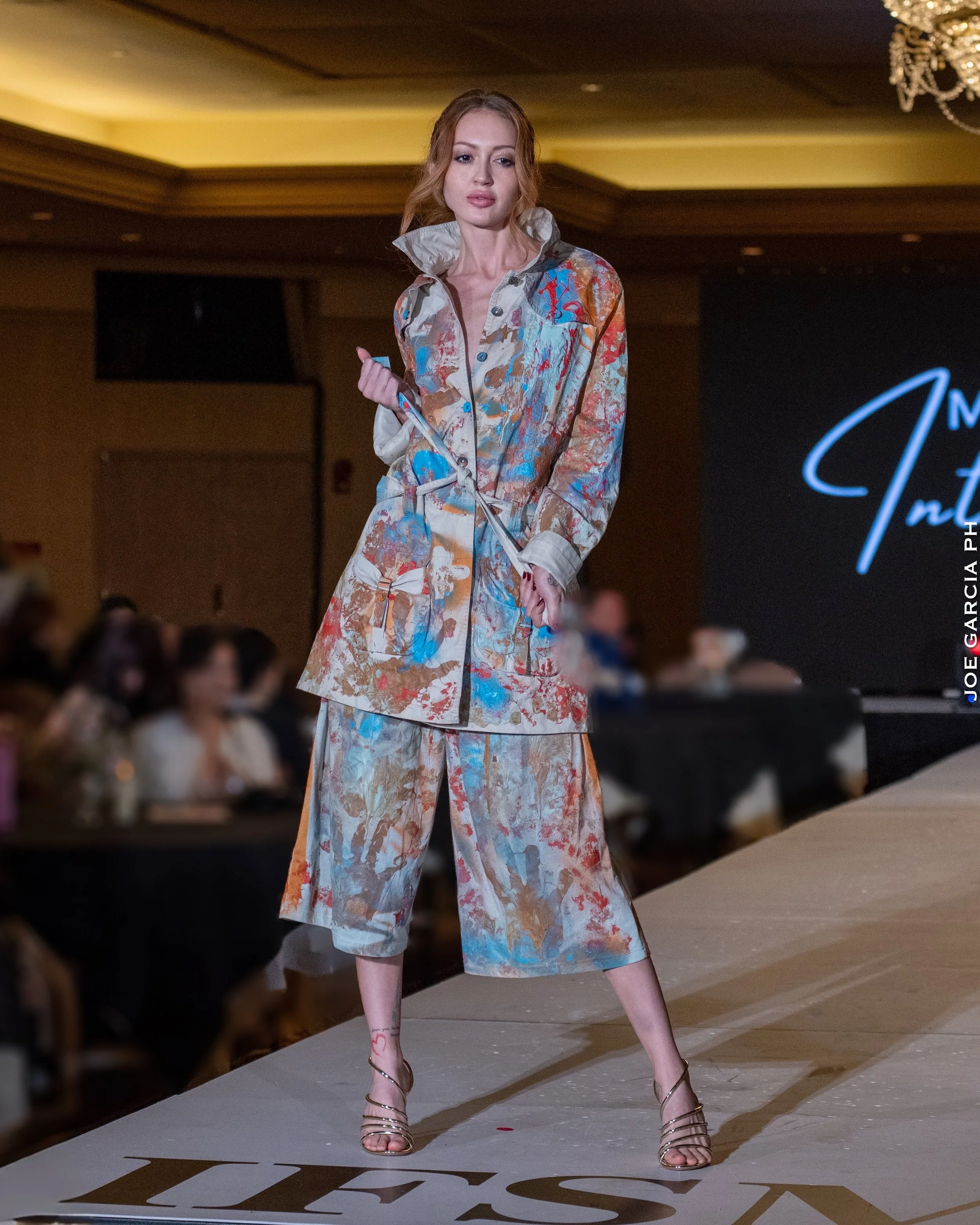 A fashion model walking on a runway at a fashion show, wearing a colorful, abstract-patterned outfit consisting of a long-sleeve jacket and wide-legged pants. The background shows an audience and a large screen with text.