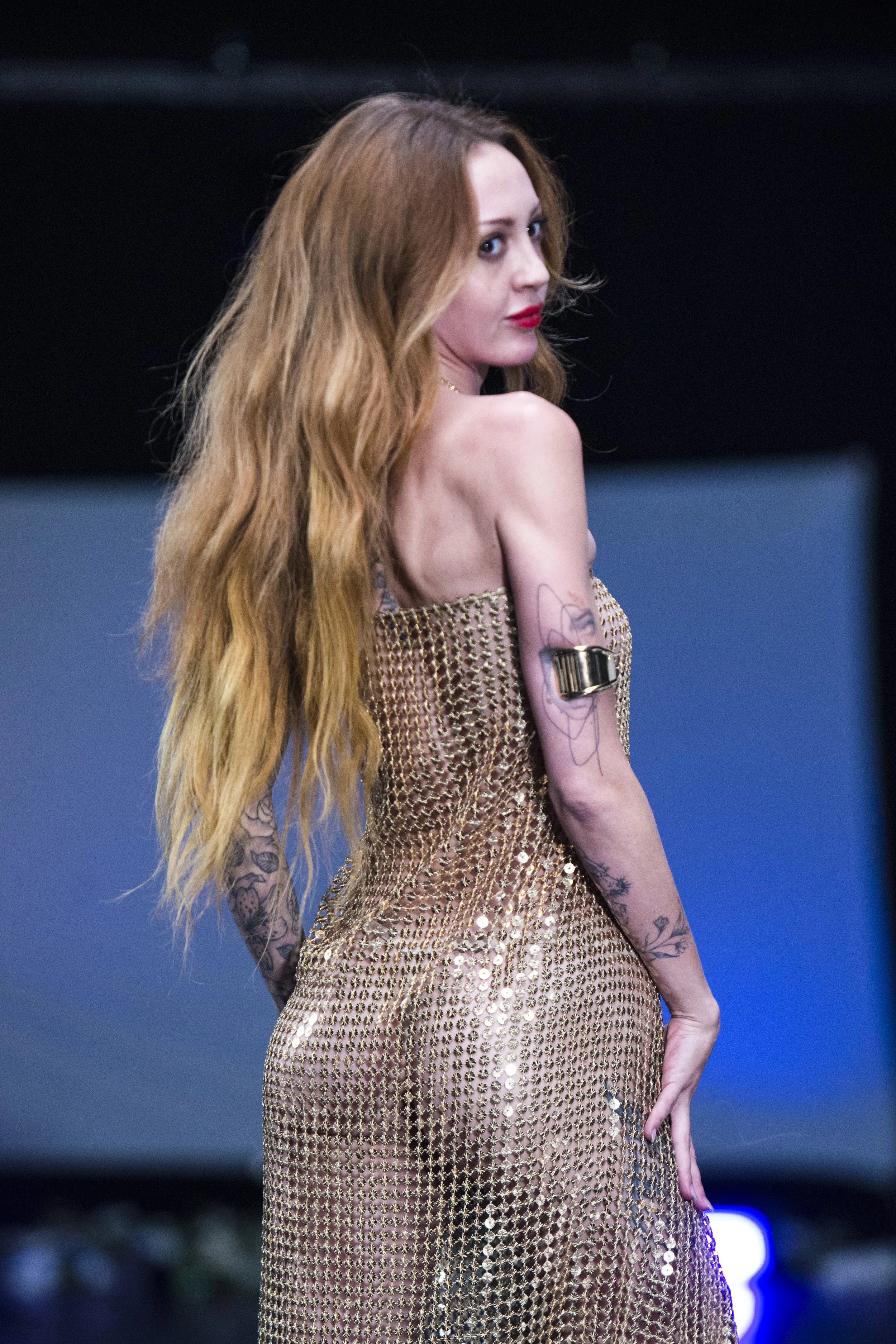 A woman with long red hair posing in a shiny, gold, see-through dress with tattoos on her arms.