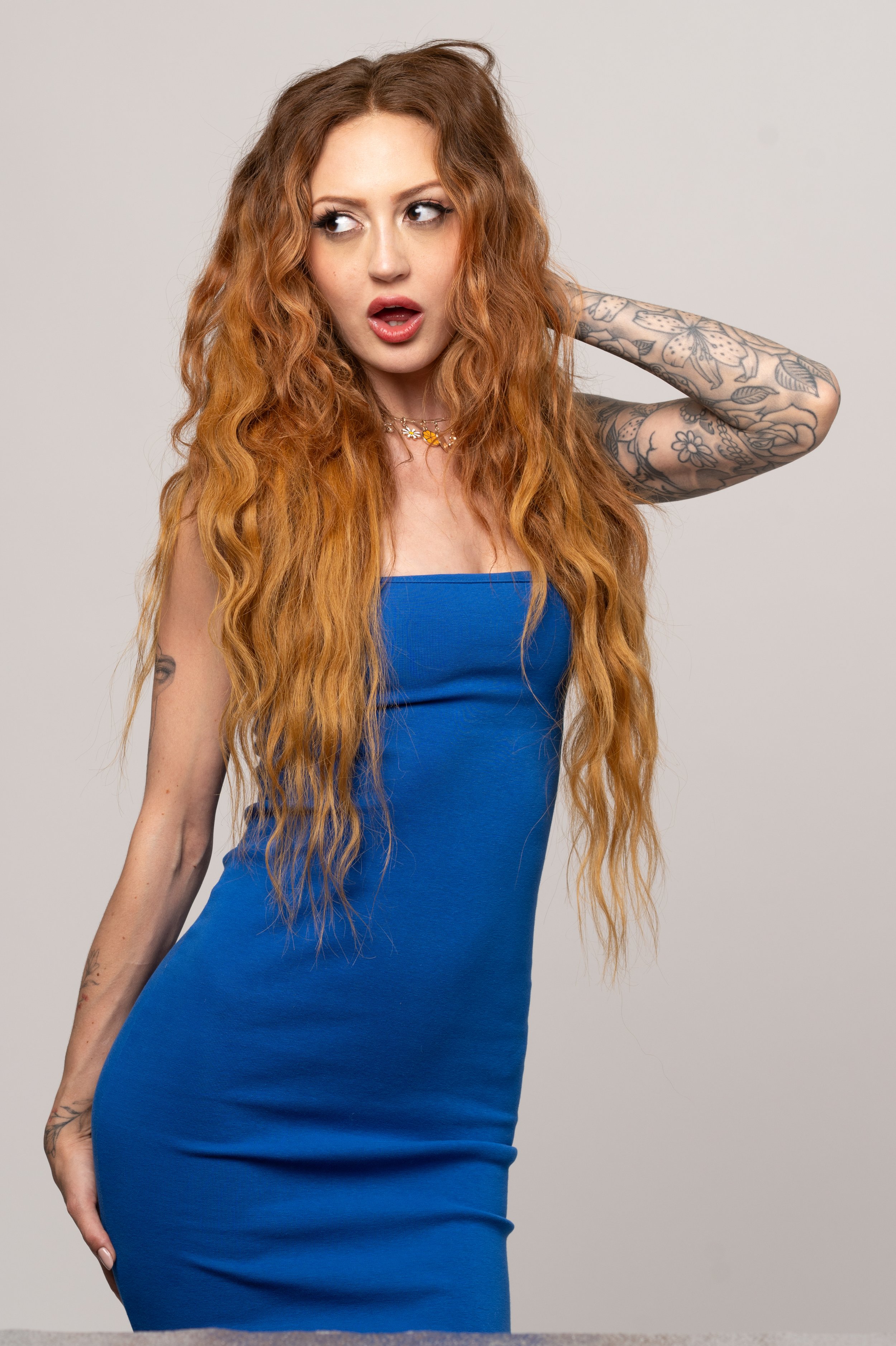 A woman with long red hair wearing a blue strapless dress, with tattoos on her arms, looking to the side with a surprised expression.