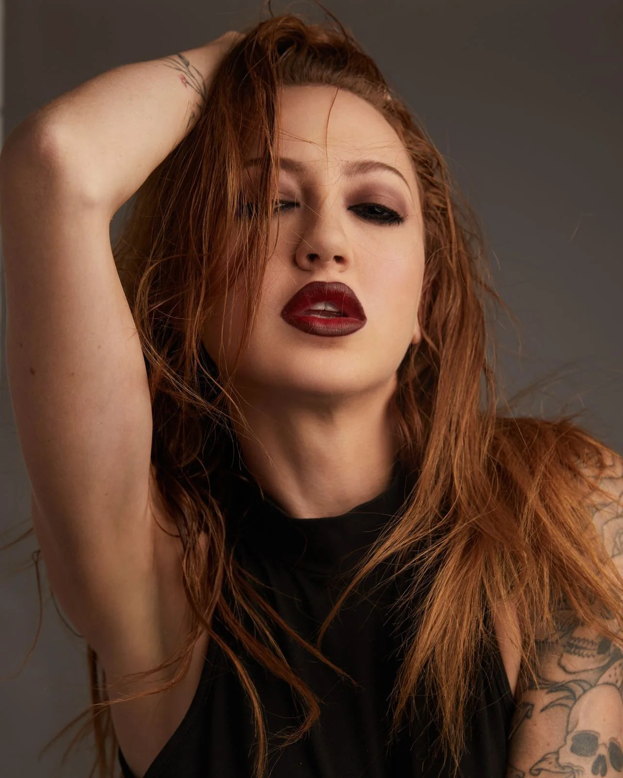 Portrait of a woman with red hair, dark lipstick, and tattoos, posing with one hand in her hair against a gray background.
