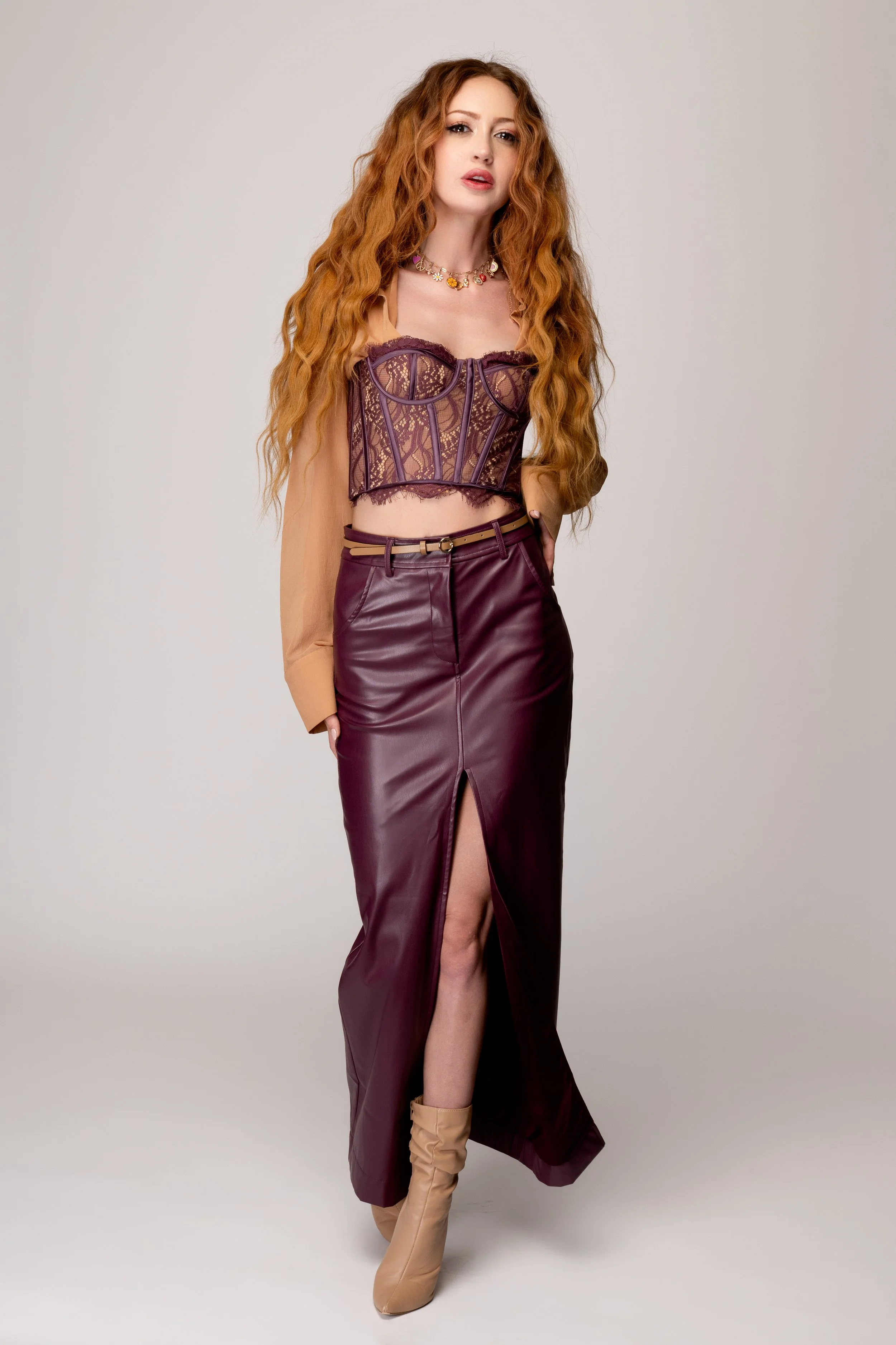 A woman with long, wavy red hair wearing a sheer brown lace crop top, a maroon leather skirt with a front slit, beige ankle boots, and a floral necklace, standing against a plain background.