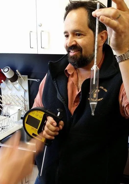A man in a lab setting holding a digital thermometer and a glass graduated cylinder with liquid, monitoring a scientific experiment.