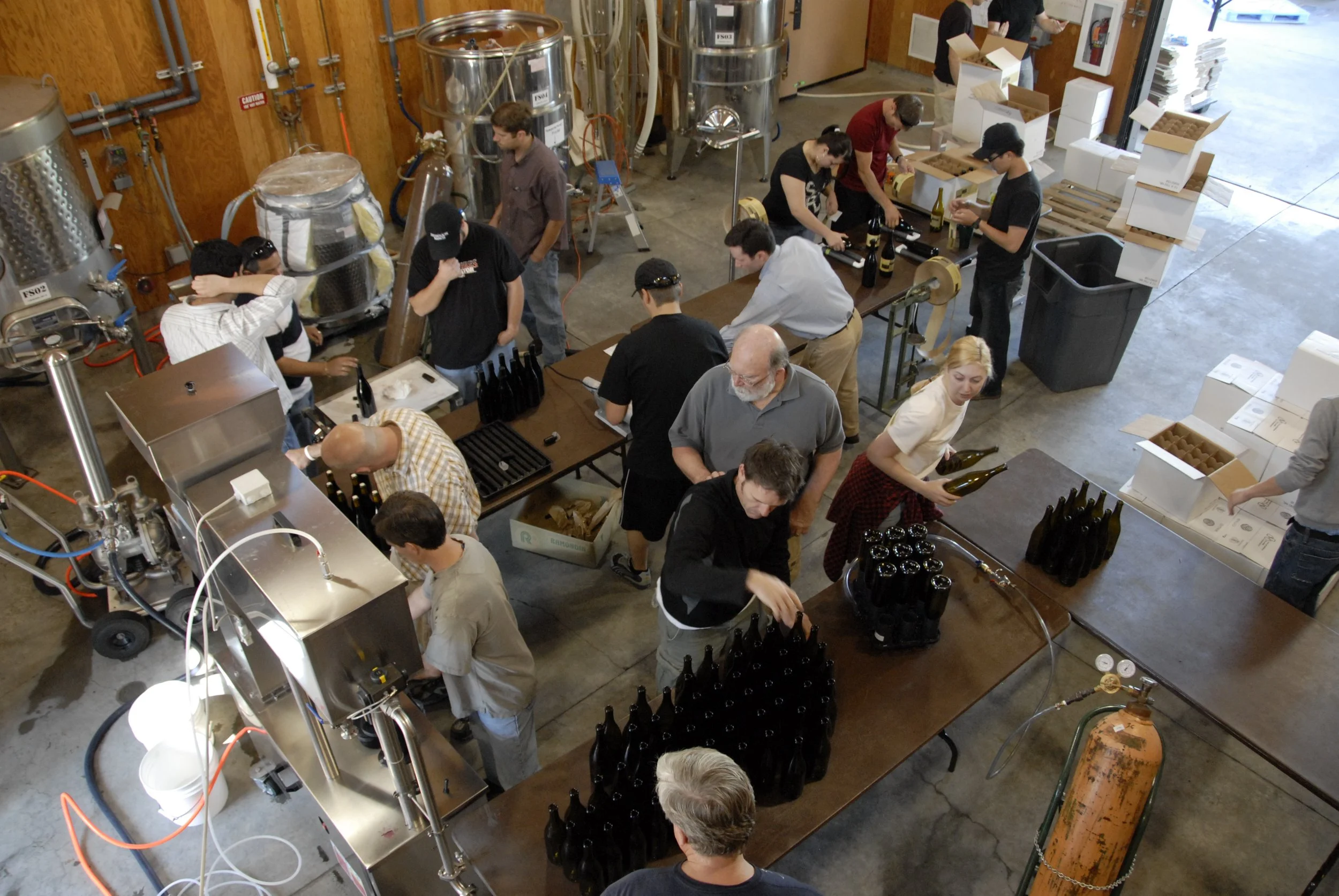 Overhead view of a brewery workshop with people working at tables. Several bottles and brewing equipment are visible, and people are inspecting, filling, and packaging bottles of beer.