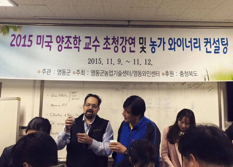 People attending a conference in a classroom with a banner above, showing two men in conversation, one holding a small cup, and others in the background