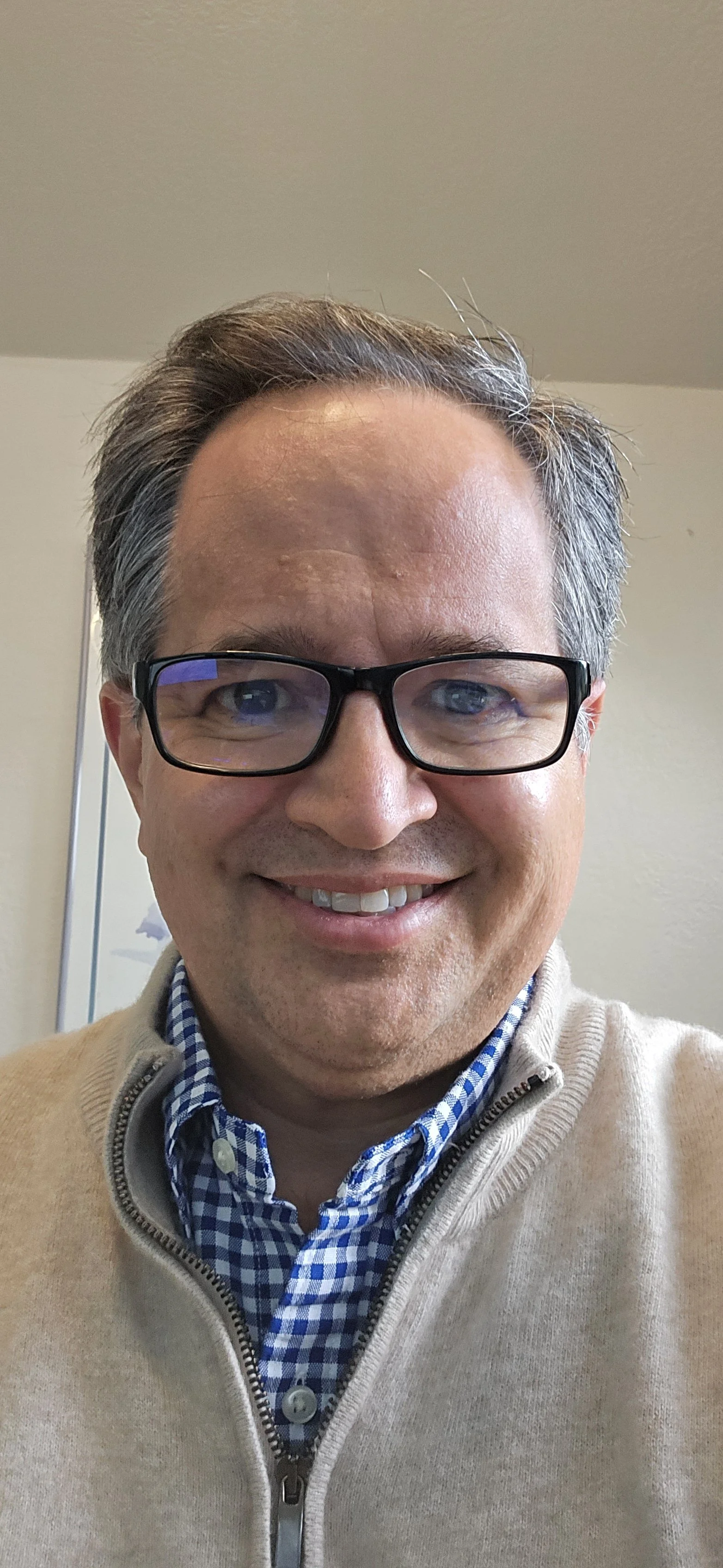 Close-up of a smiling man wearing glasses, a beige zip-up sweater, and a blue checkered shirt in an indoor setting.