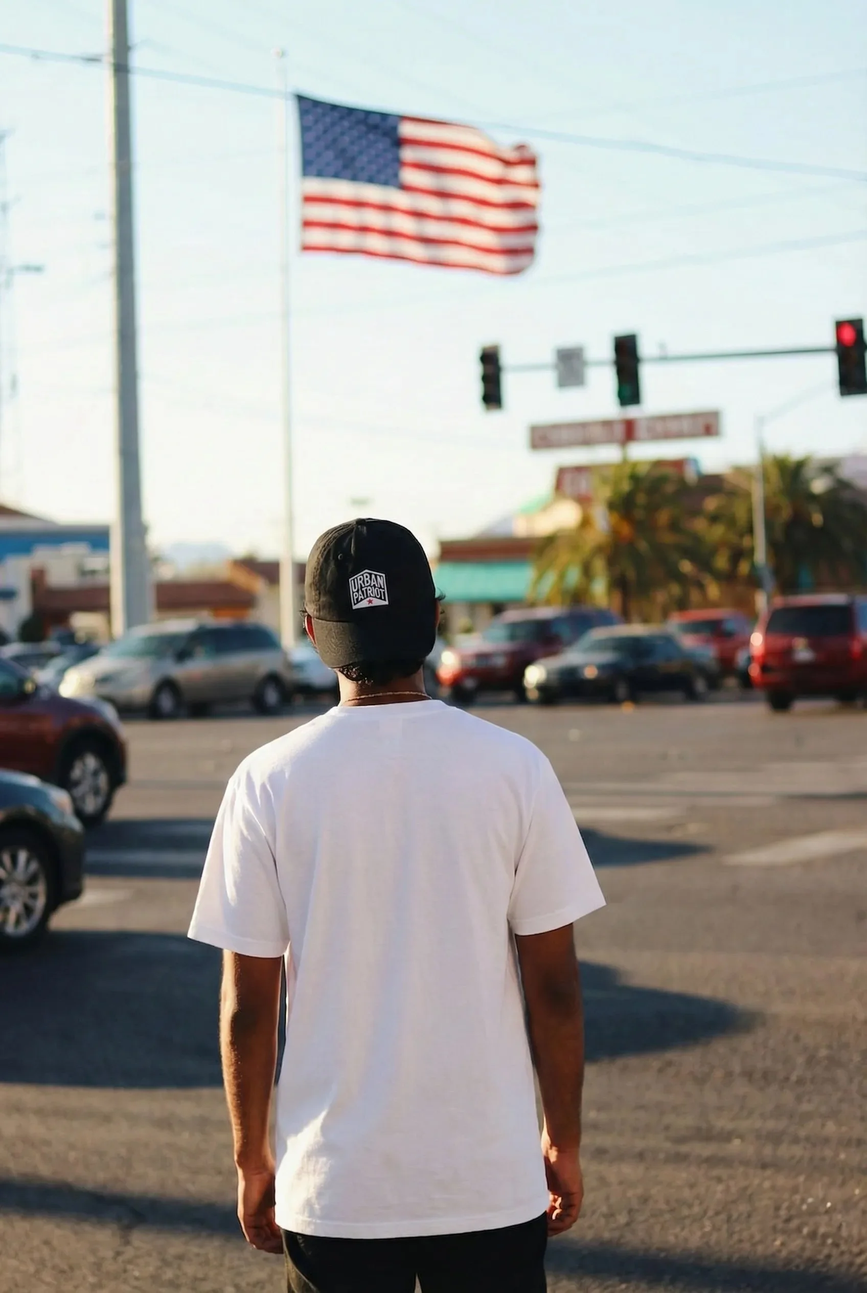 A person wearing a black cap and white shirt walking in a parking lot with multiple cars and an American flag in the background.