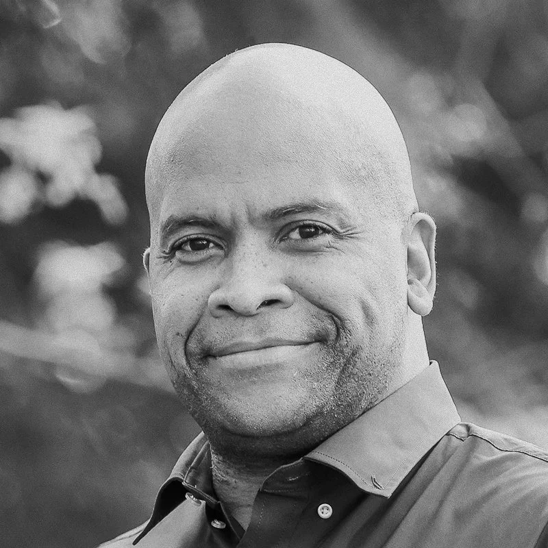 A black and white photo of a smiling man with a shaved head and wearing a collared shirt, outdoors with blurred foliage in the background.
