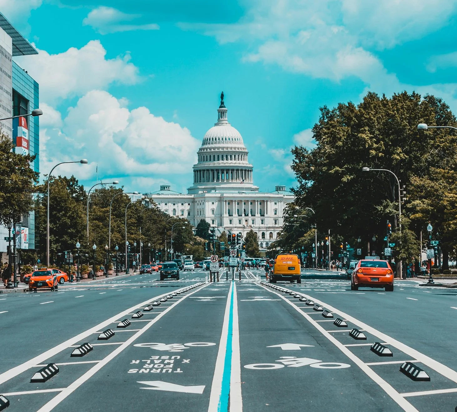 View of the U.S. Capitol building in Washington, D.C., with a street in the foreground featuring designated bike lanes and traffic. Cars and pedestrians are present, with trees lining the street and a partly cloudy sky above.