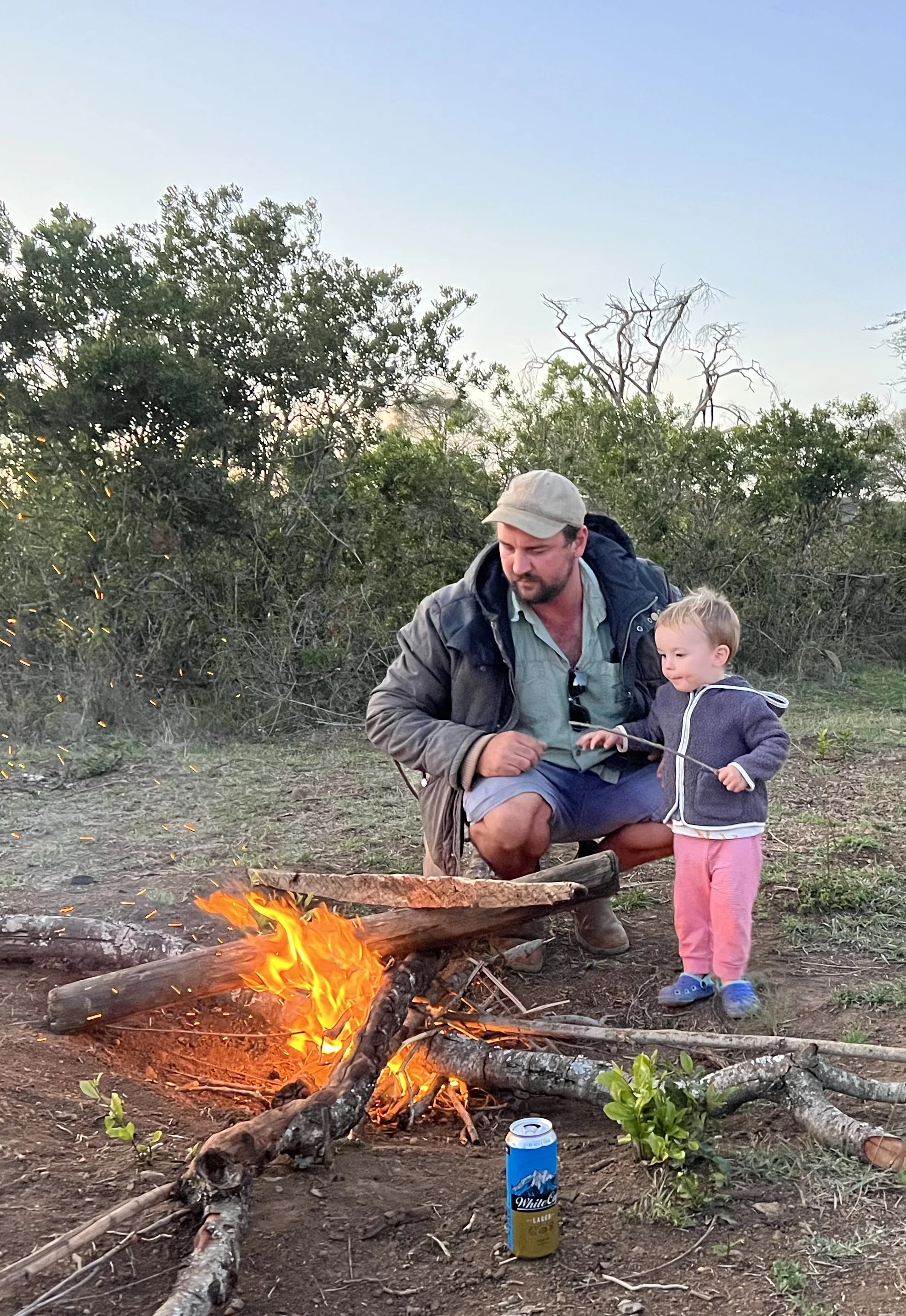 A man and a young girl sitting by a campfire outdoors with trees in the background. The girl is holding a stick over the fire, and there is a can of White Claw hard seltzer on the ground nearby.
