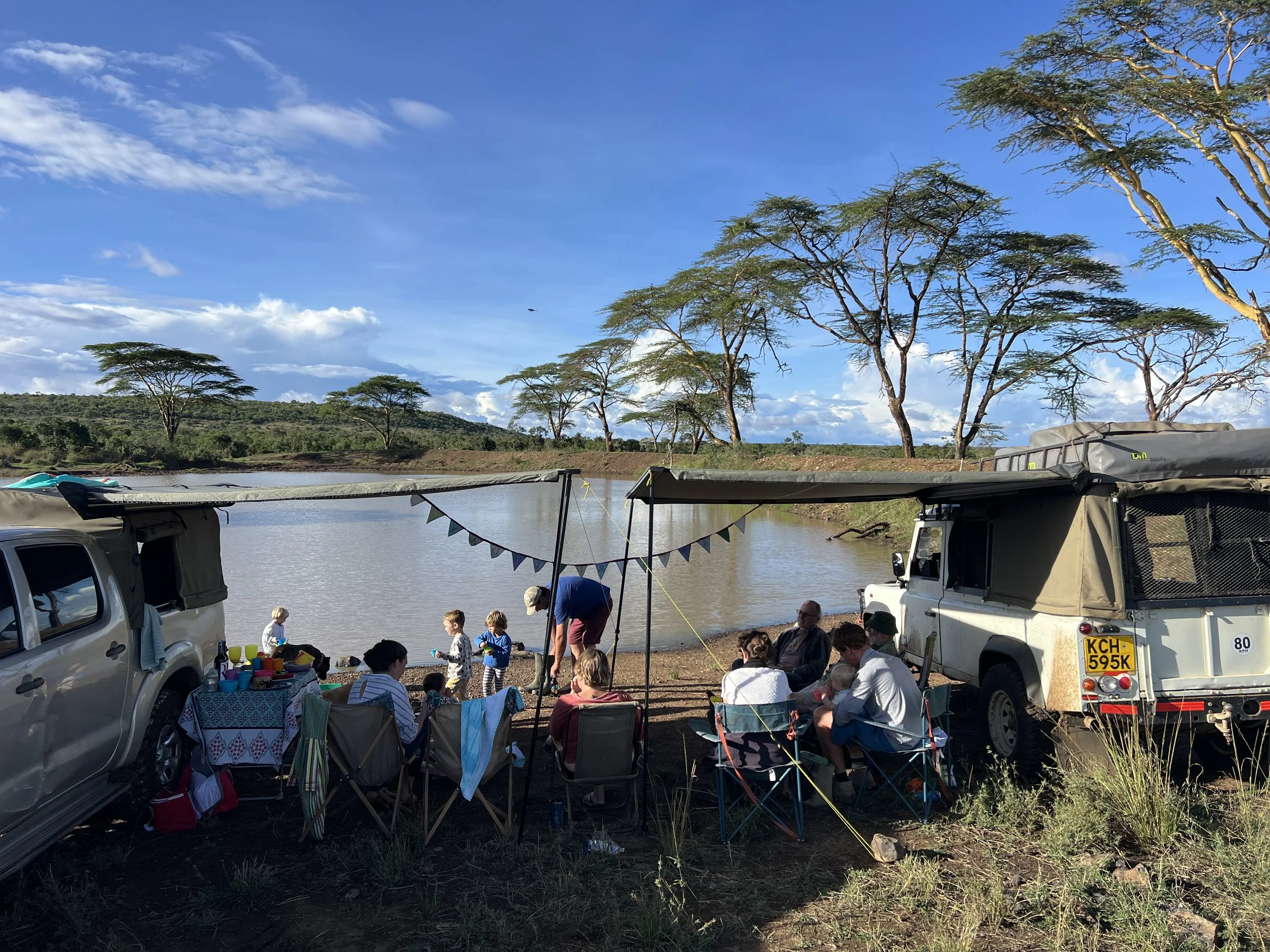 People sitting and standing around a campsite by a river with trees and a blue sky in the background.