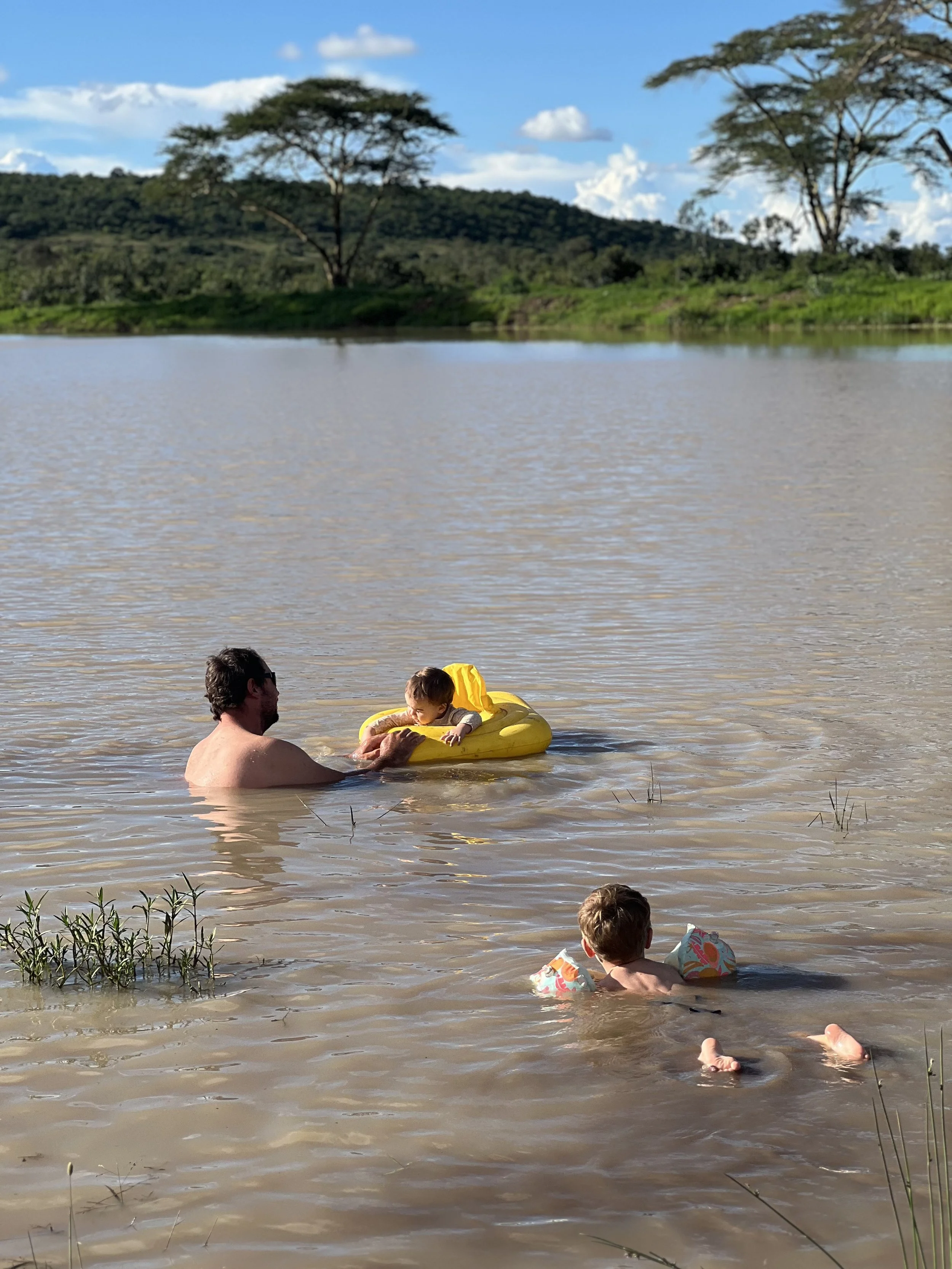 A man swimming in a lake with two children, one in a yellow inflatable boat and the other with floaties, surrounded by a river, green trees, and hills under a blue sky with clouds.