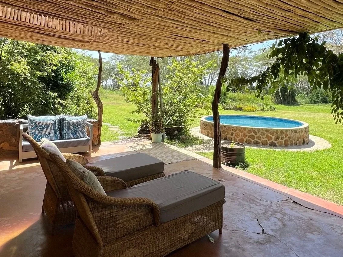 Outdoor patio with wicker chairs and a sofa, overlooking a grassy yard with a small stone hot tub and lush greenery under a thatched roof.