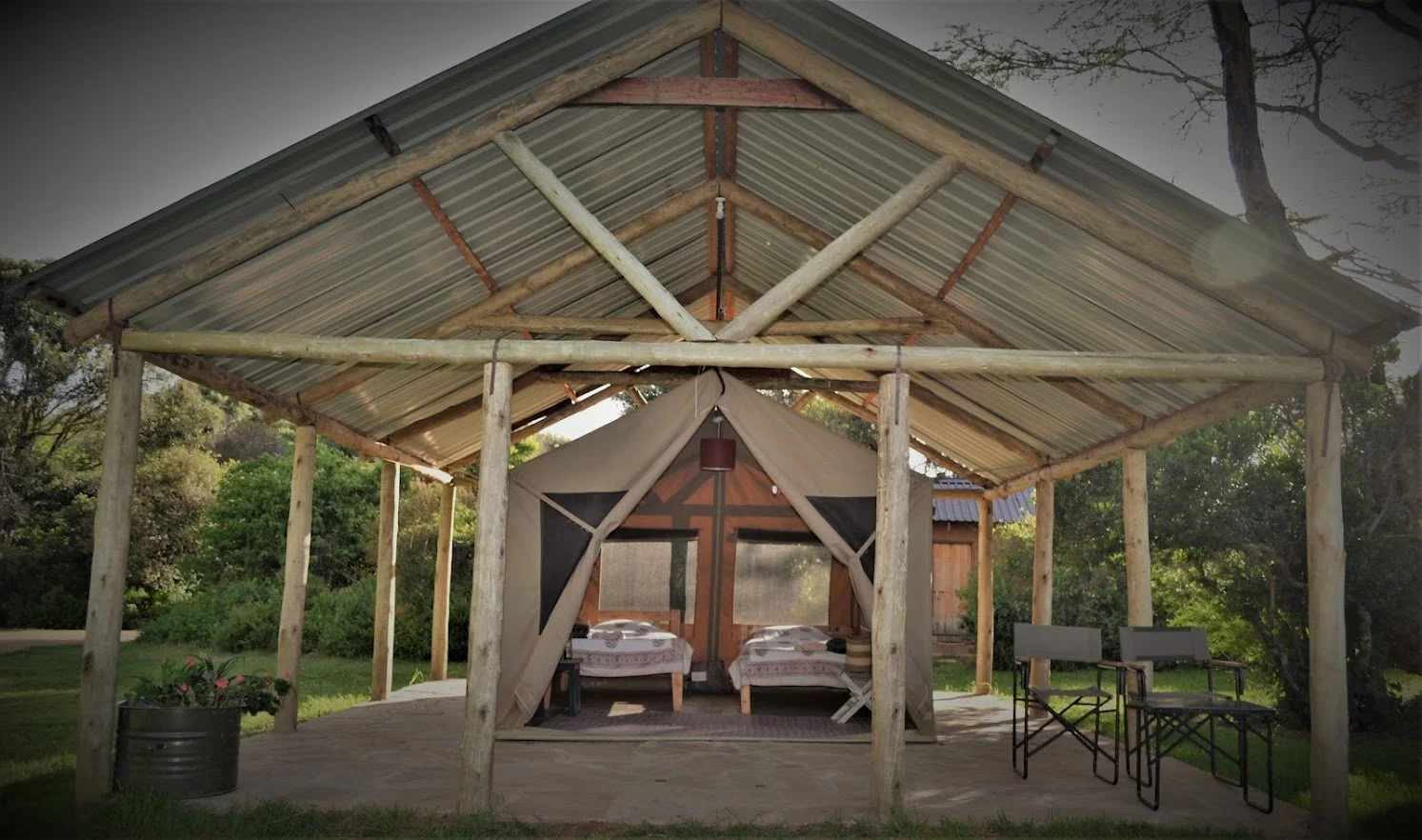 A rustic outdoor cabin with a metal roof, two beds inside, and outdoor seating with two chairs and small tables, surrounded by greenery.