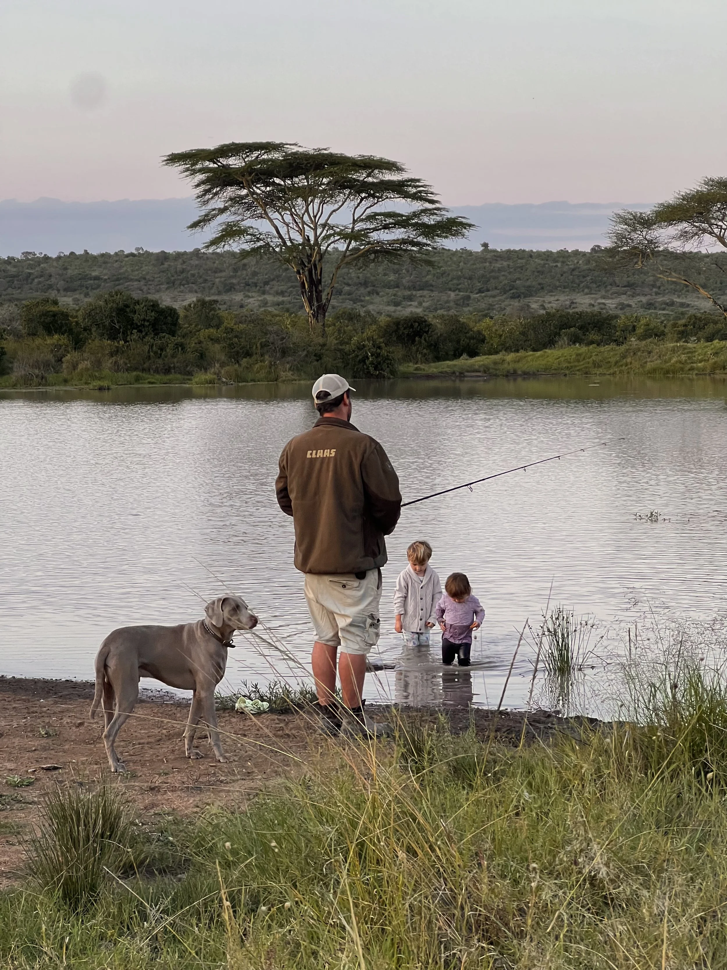 A man with a dog and two children fishing at a lake with trees in the background