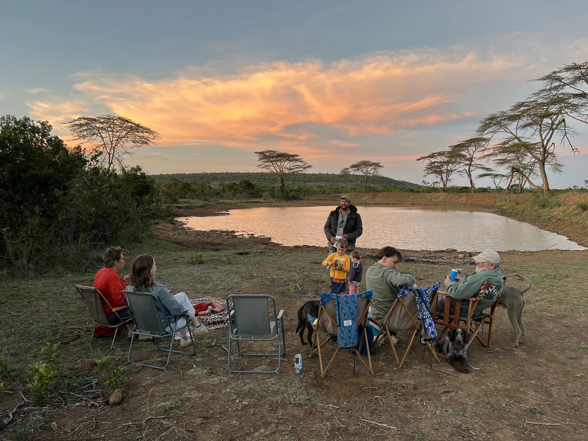 A group of seven people, four adults and three children, gathered outdoors by a small lake during sunset. They are sitting in camping chairs and on the ground, with some dogs nearby. The sky is partly cloudy with pink and orange hues, and the landsca