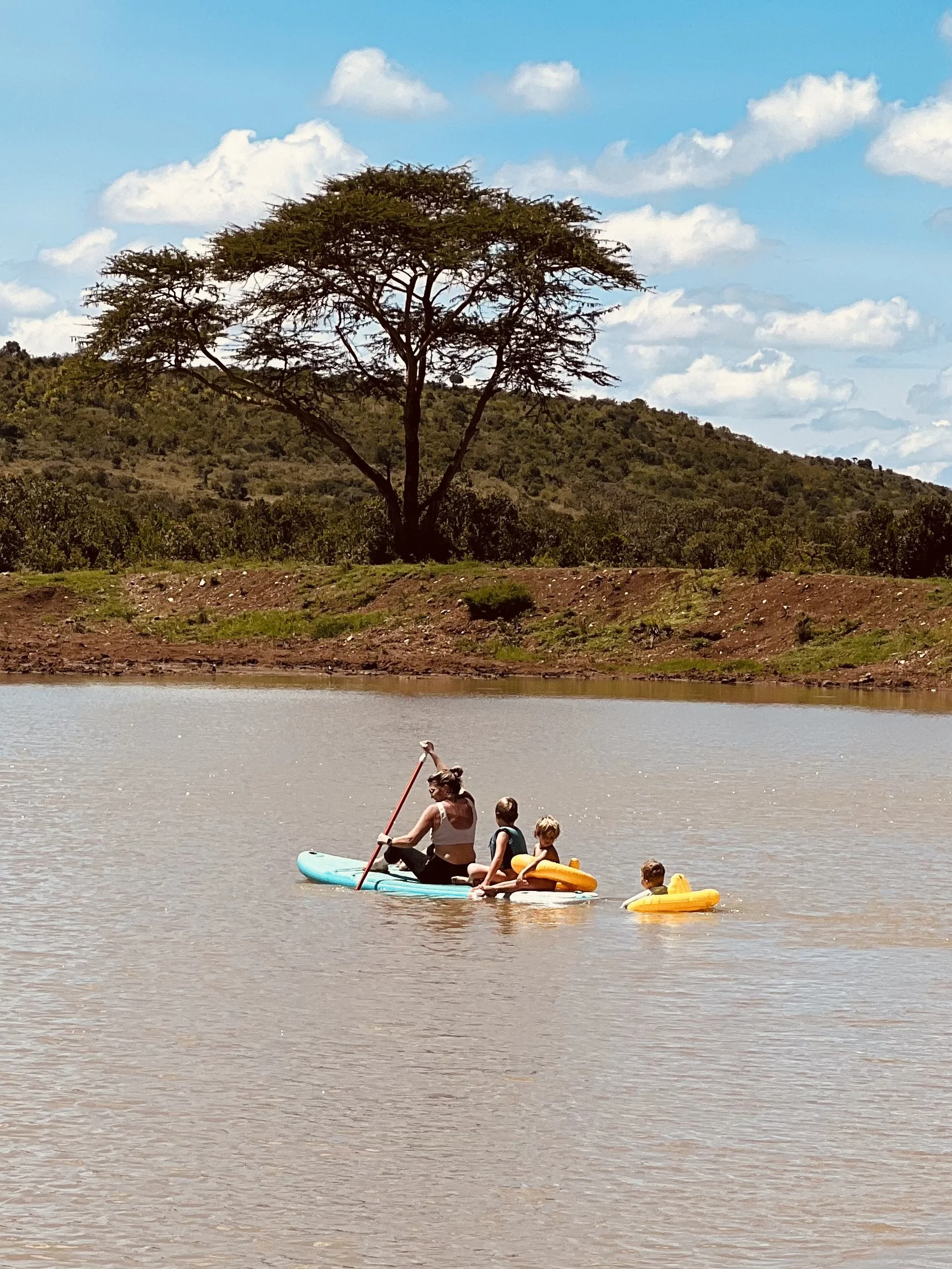 A woman and three children paddle and float on a body of water, with a large tree and hilly landscape in the background, under a partly cloudy sky.