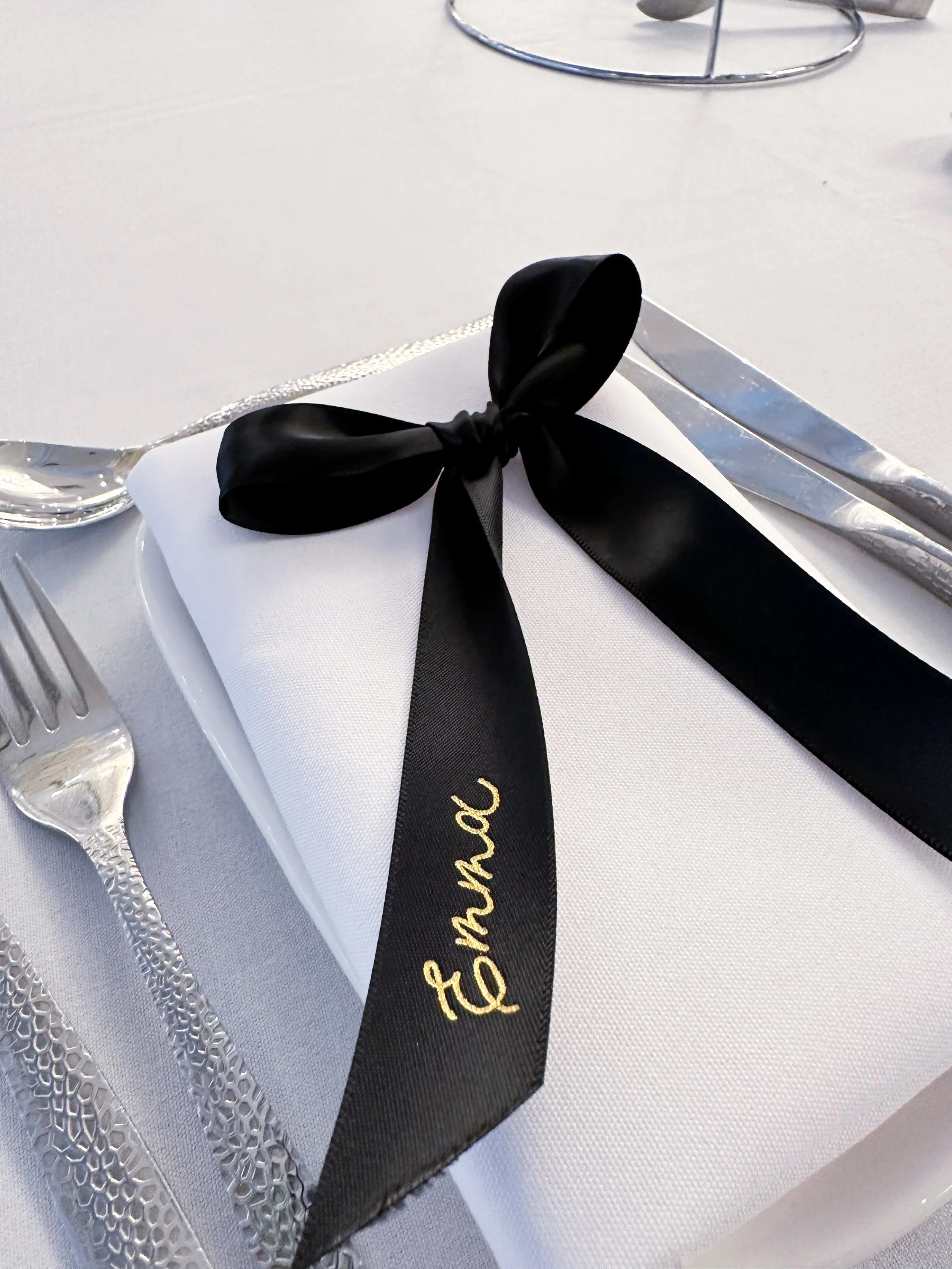 Elegant table setting with black ribbon tied in a bow on a white napkin, inscribed with 'bride' in gold, surrounded by silver cutlery and a glass.