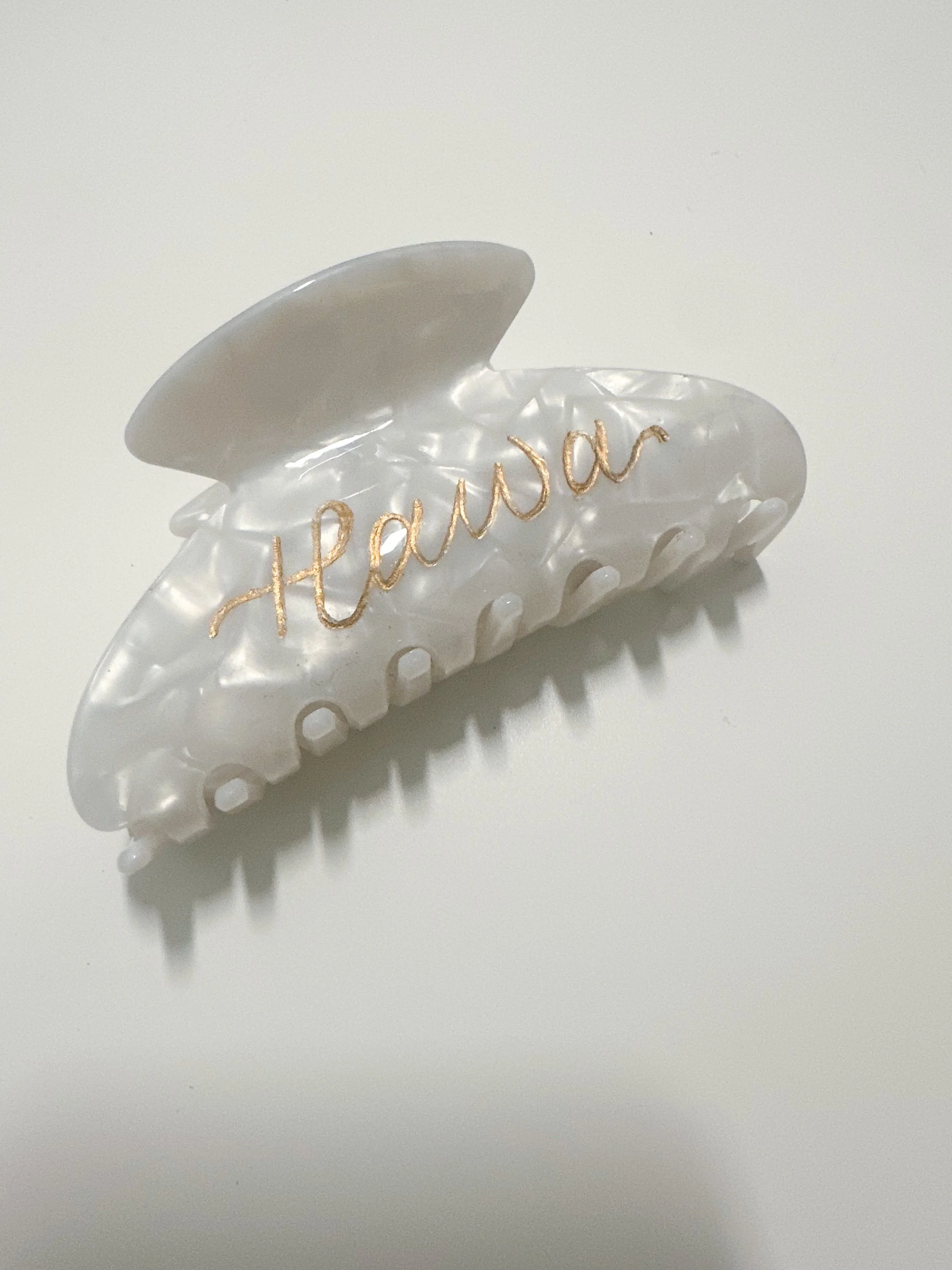 A white plastic hair clip with a pearl-like texture, decorated with the word 'Hearwar' written in gold cursive.