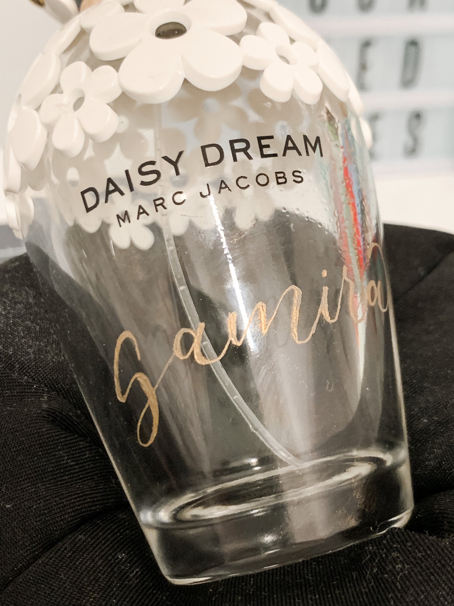 A clear glass tumbler with decorative white flowers near the top and gold lettering that reads 'gamine.' The glass is labeled 'Daisy Dream Marc Jacobs'.