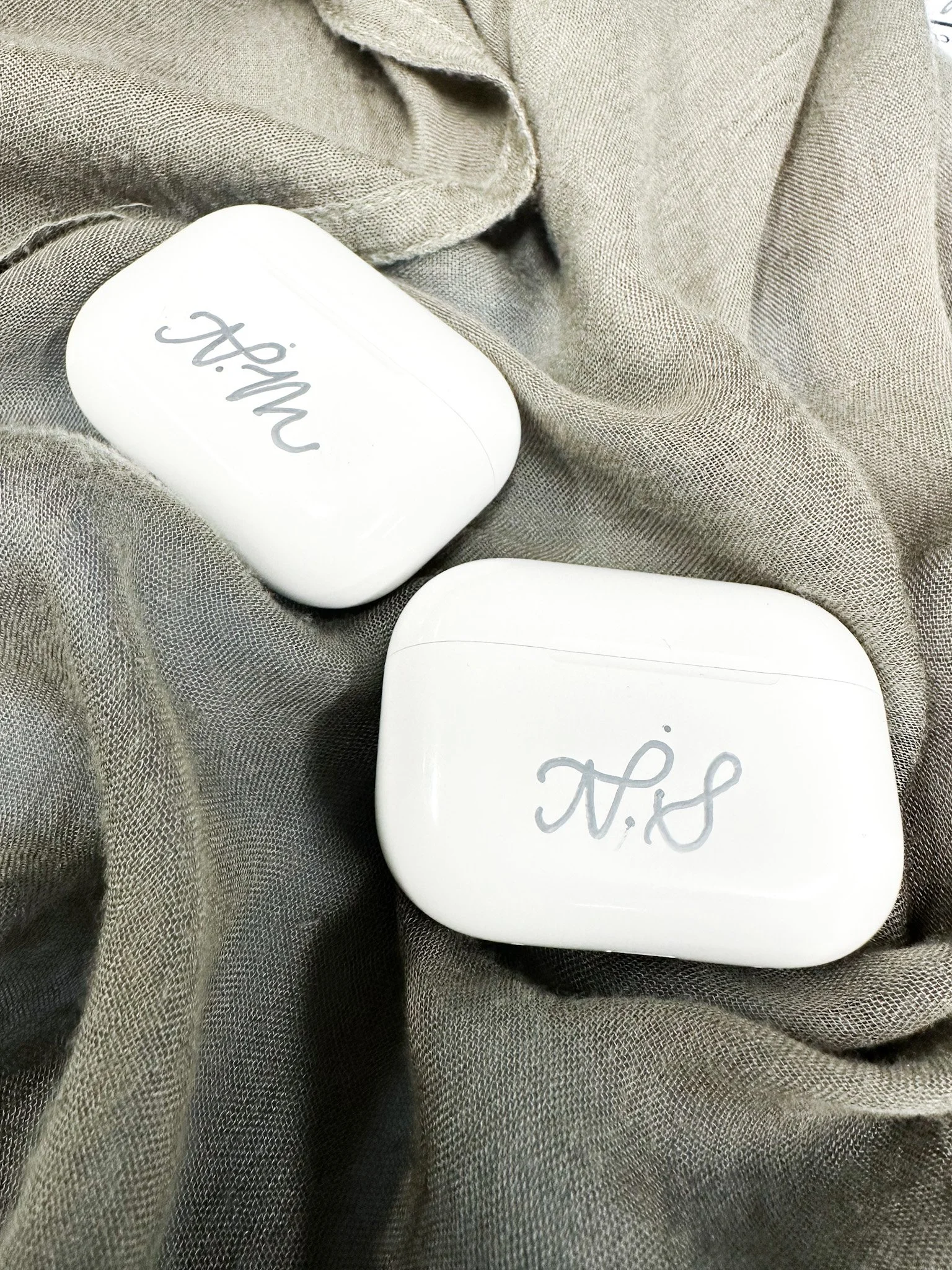 A pair of white AirPods cases with handwritten labels. One labeled 'Mom' and the other labeled 'S.' They are resting on a soft, textured beige cloth.