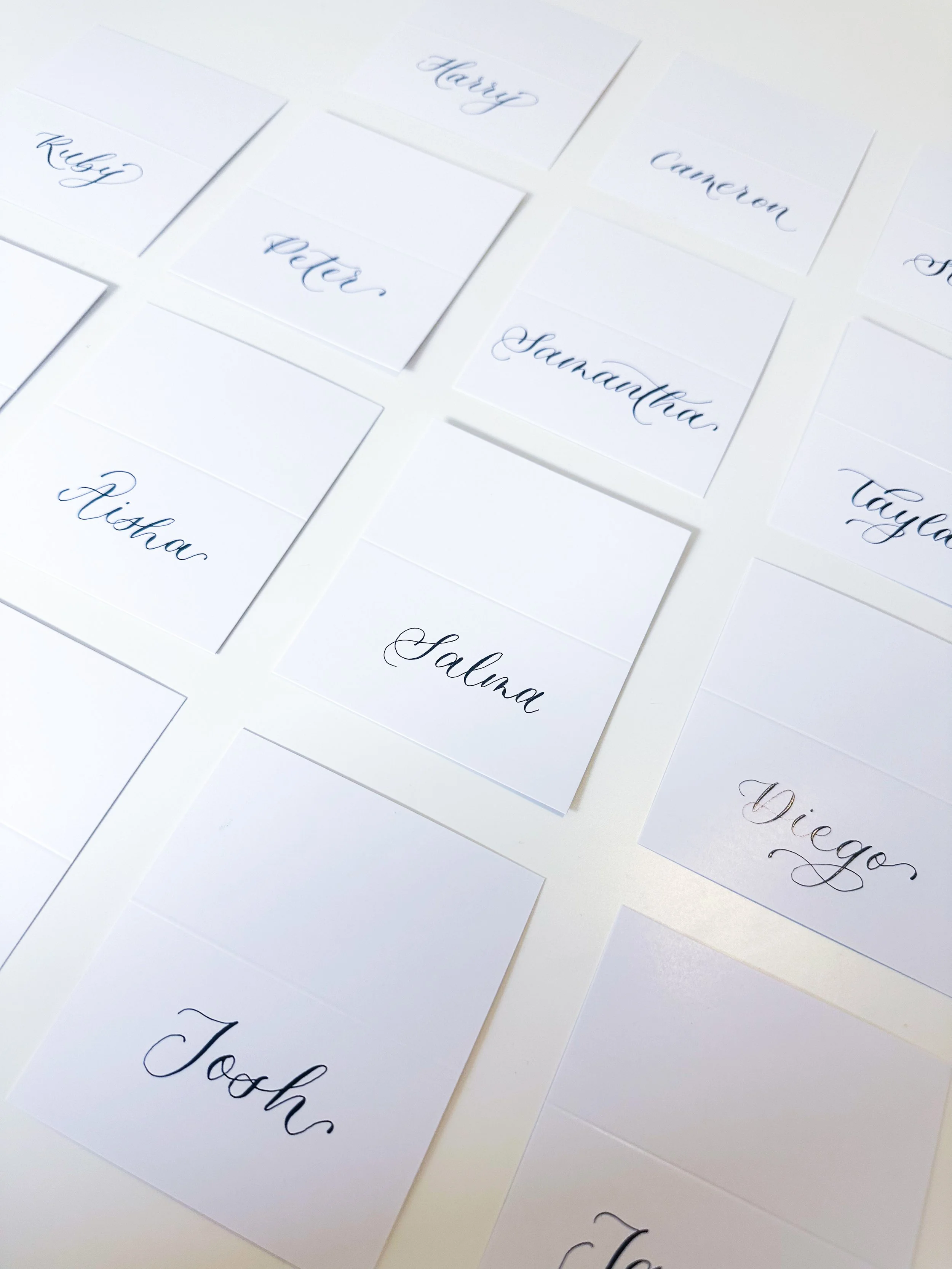 Strips of paper with handwritten names in black and blue ink, arranged on a white surface. Names include Ruby, Heather, Cameron, Peter, Savannah, Bishop, Salma, Diego, Josh, Taysia.