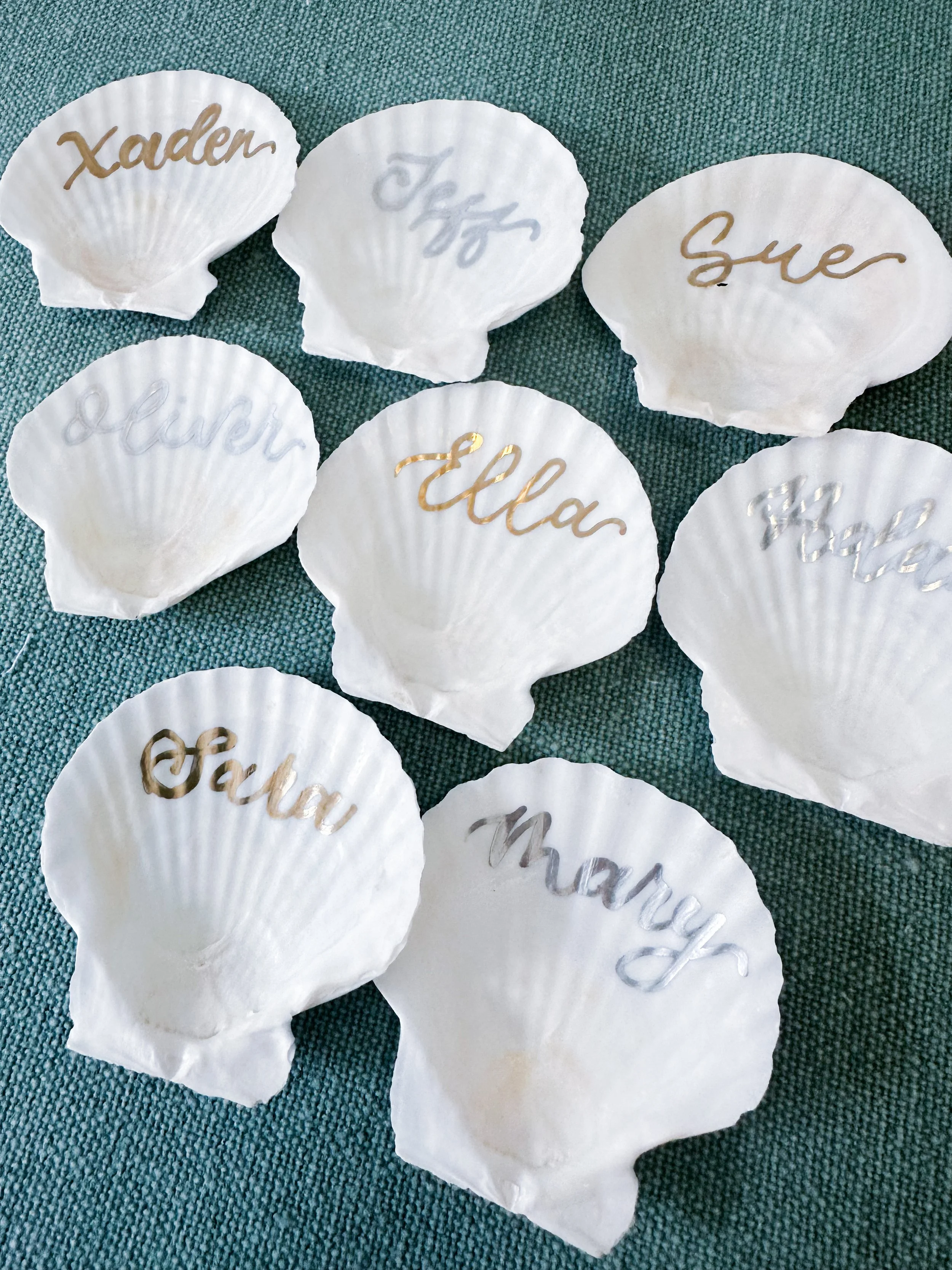 White seashells with names written in gold or silver cursive, including Xaden, Jyp, Syer, Levi, Ella, The, Marcy, and Saga, spread on a teal fabric background.