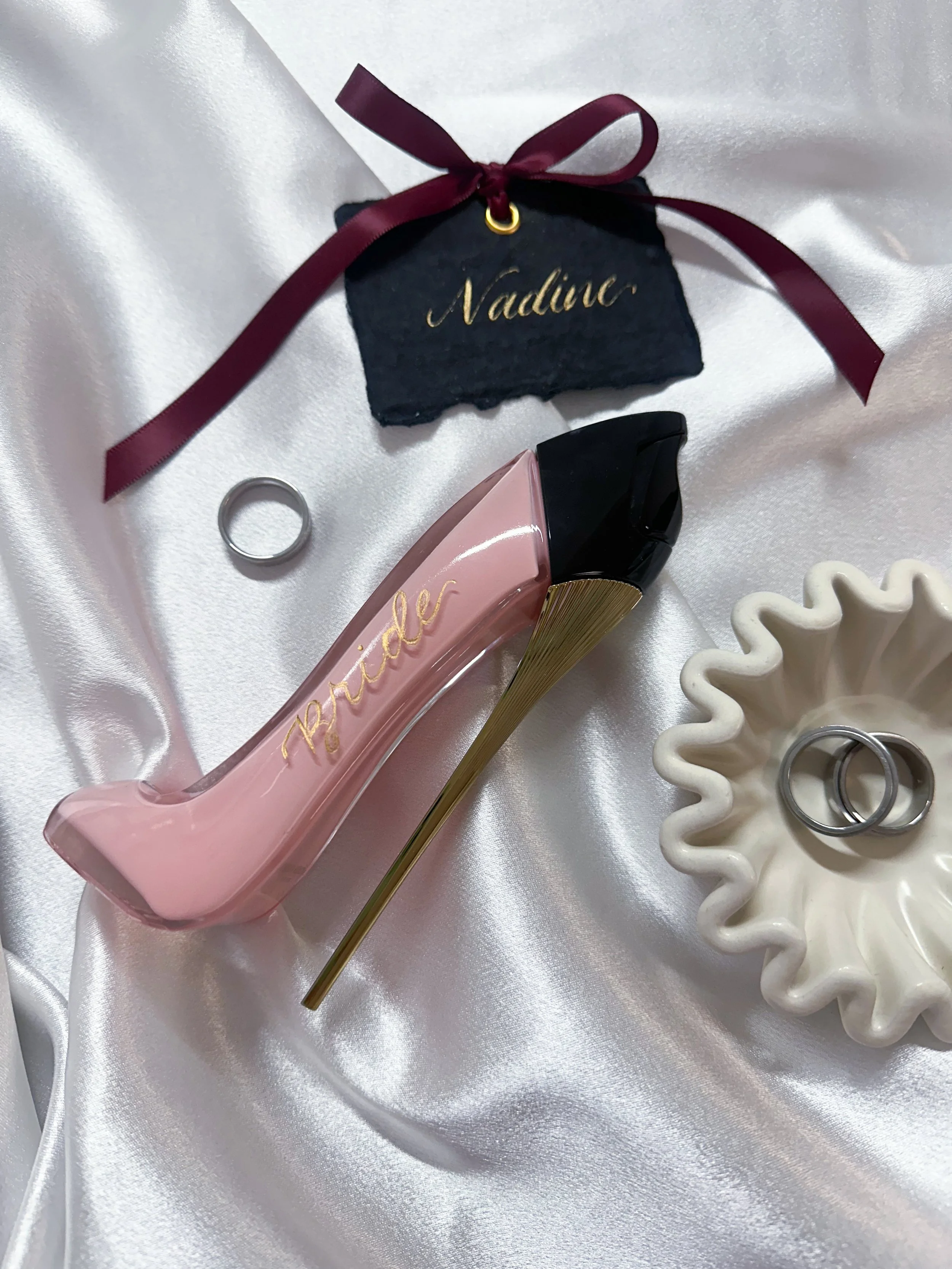 Pink high heel shoe-shaped perfume bottle with the name 'Prada' written on it, placed on white satin fabric amid rings, a black slate tag with gold writing 'Nadine' tied with a burgundy ribbon, and a cream-colored decorative dish with two rings inside.