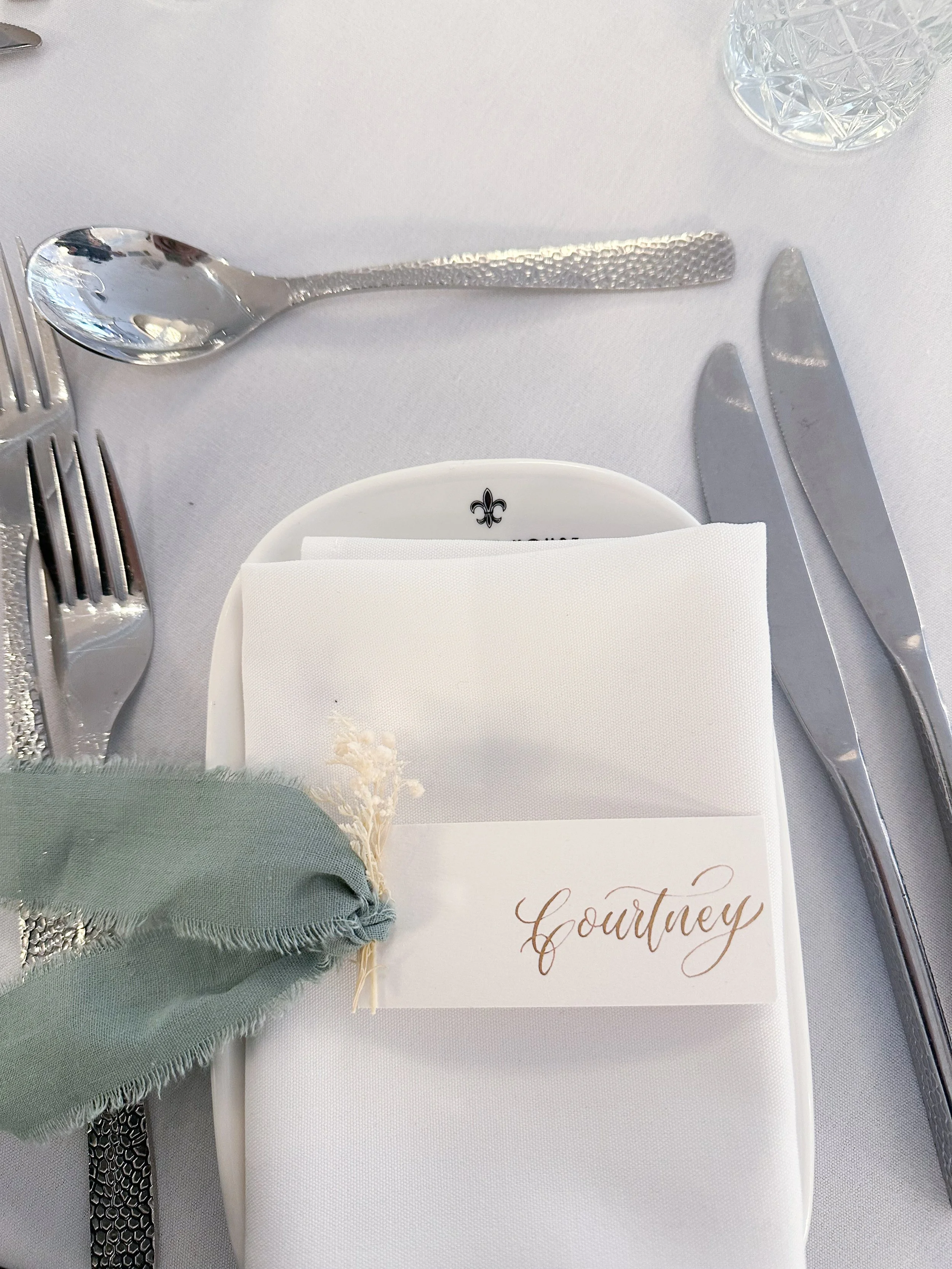 Place setting with a white plate, a folded white napkin, a green napkin tied with a silver napkin ring, and silverware including a fork, spoon, and knives, with a guest name tag reading "Courtney" on a formal dining table.