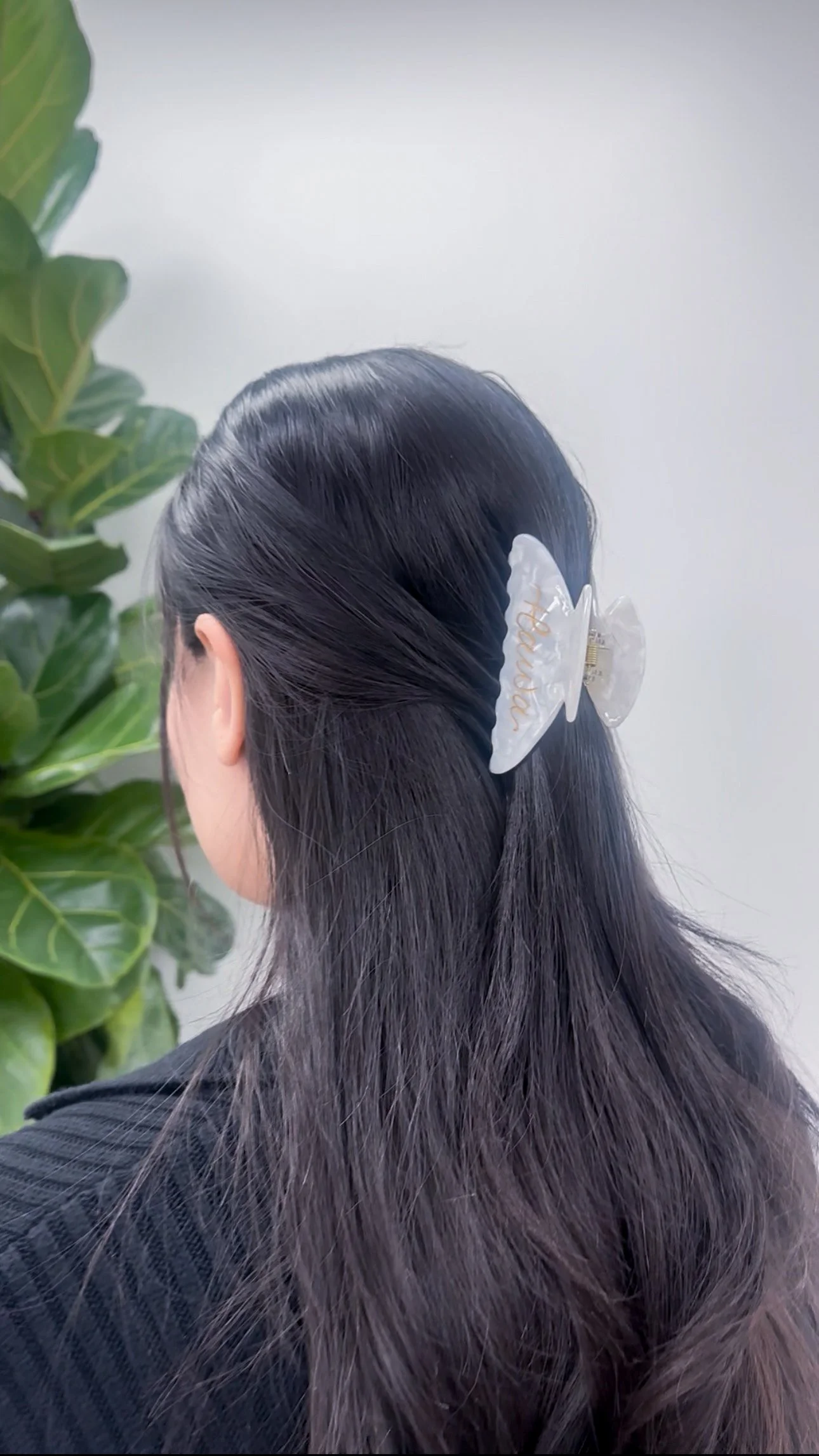 A woman with long, dark hair secured with a clear hair clip that has the word 'happiness' written in gold, standing next to a large green plant against a white wall.
