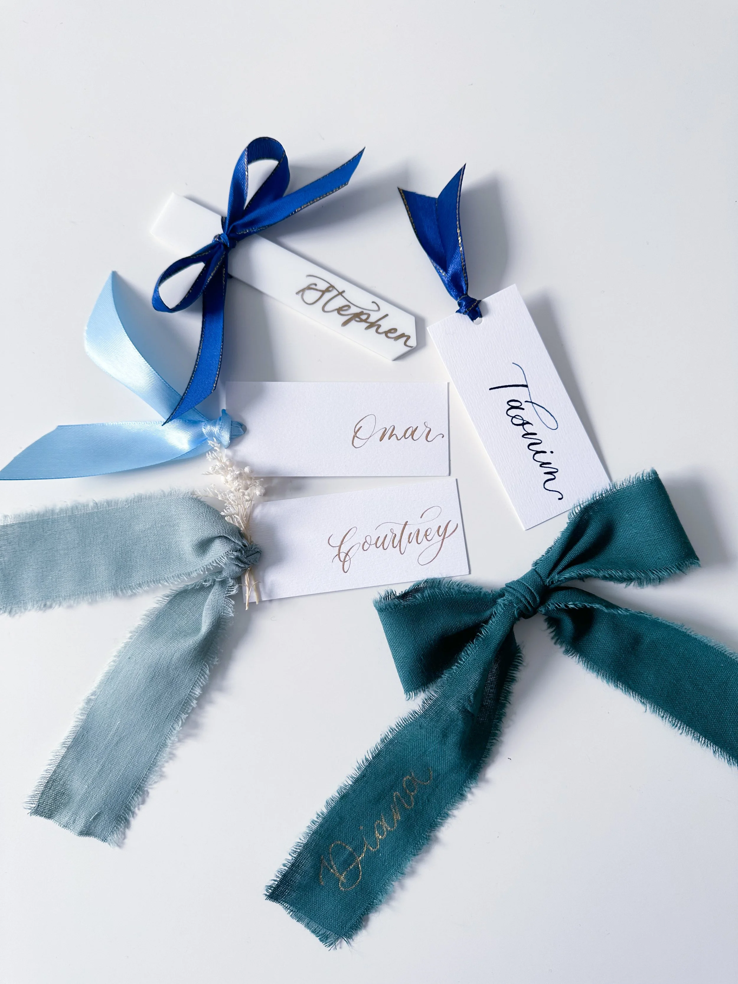 Four ribbon bookmarks with name tags attached, placed on a white surface. Each bookmark has a different color ribbon and a name written on it: Stephen, Tobias, Omar, Courtney, and Dianne.