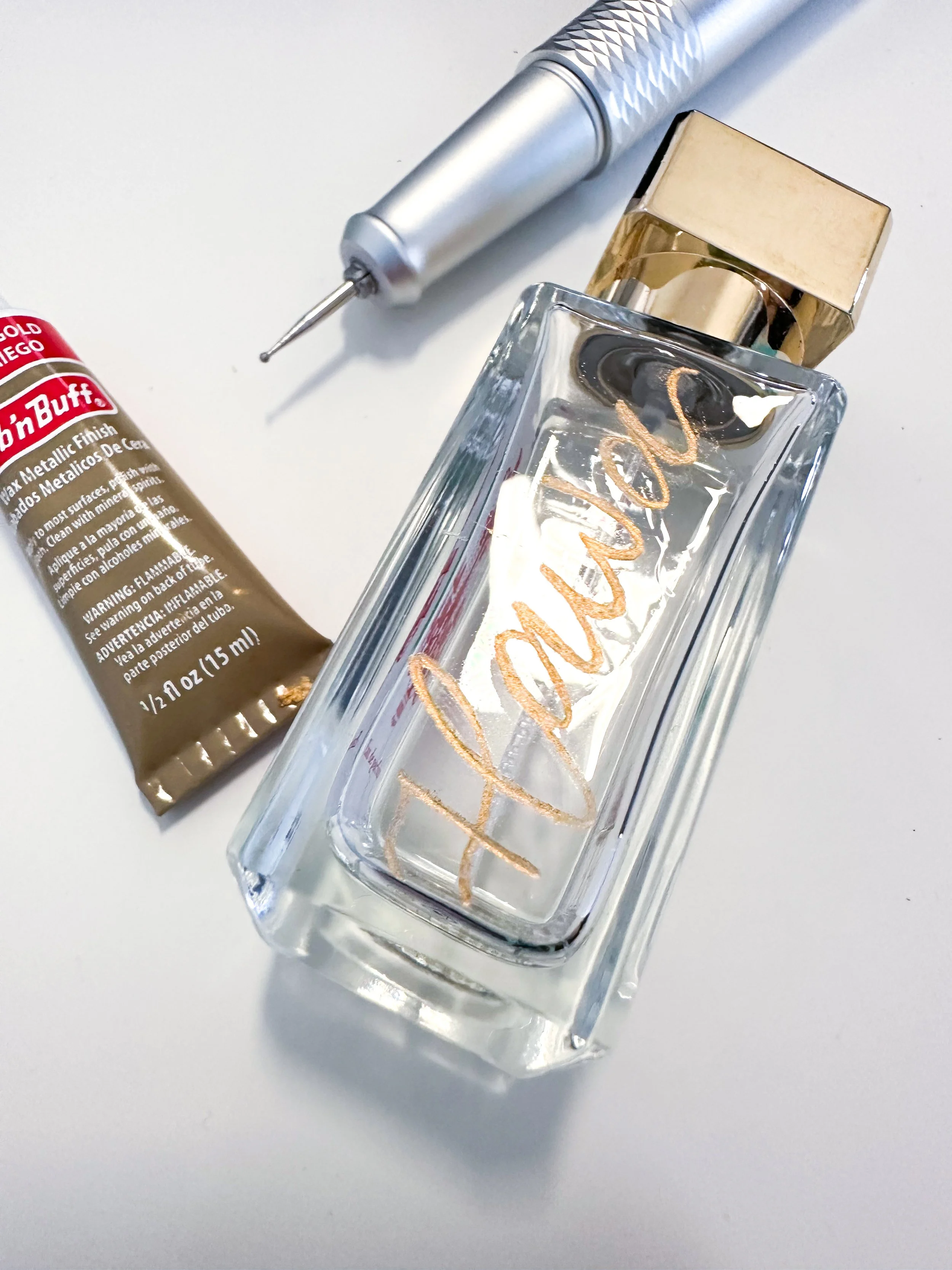 A perfume bottle with the word 'Fenty' on it, a tube of Robin Buff metallic finish paint, and a nail drill on a white surface.