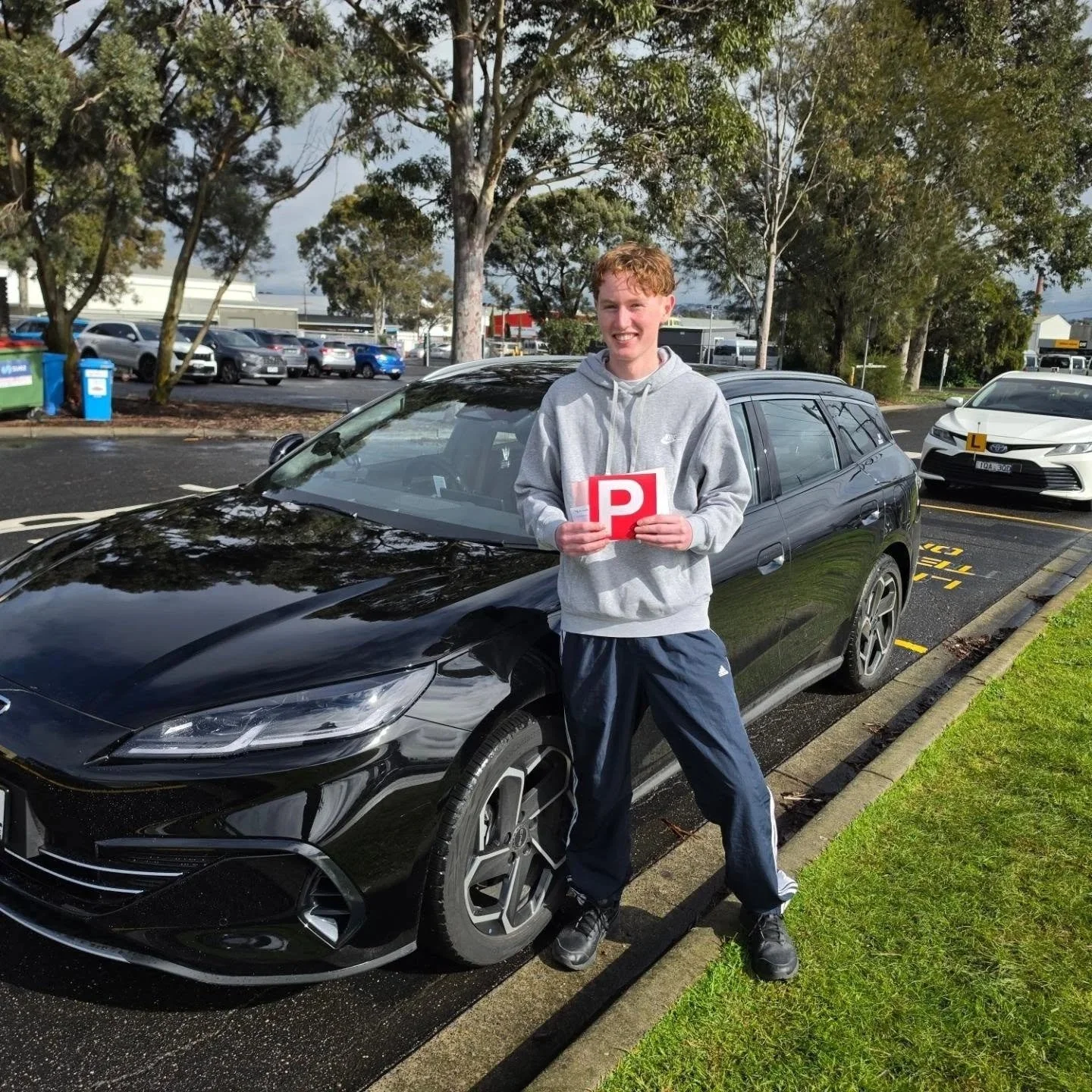 Smiling learner driver giving a thumbs-up beside a driving lesson vehicle after a driving lesson.