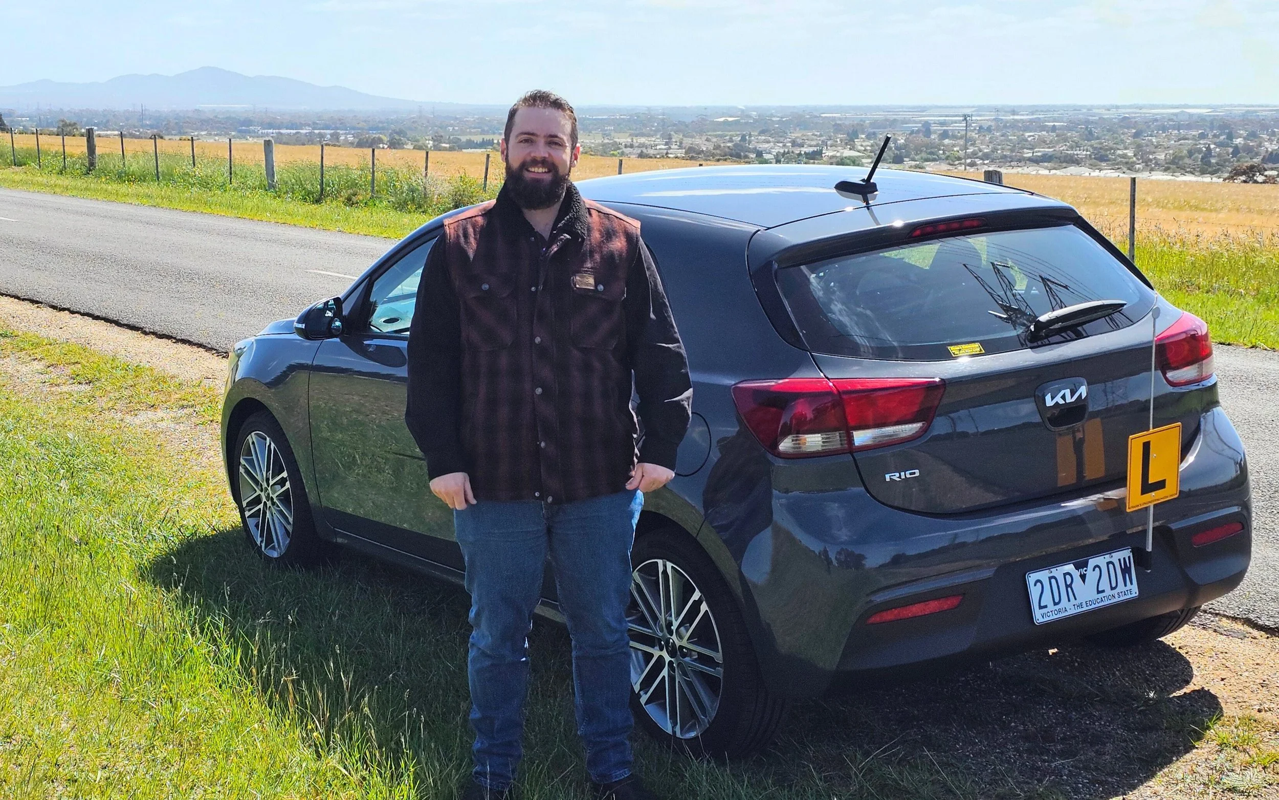 WhiteLine Driving School instructor beside a grey Kia Rio manual driving lesson vehicle on a scenic roadside.
