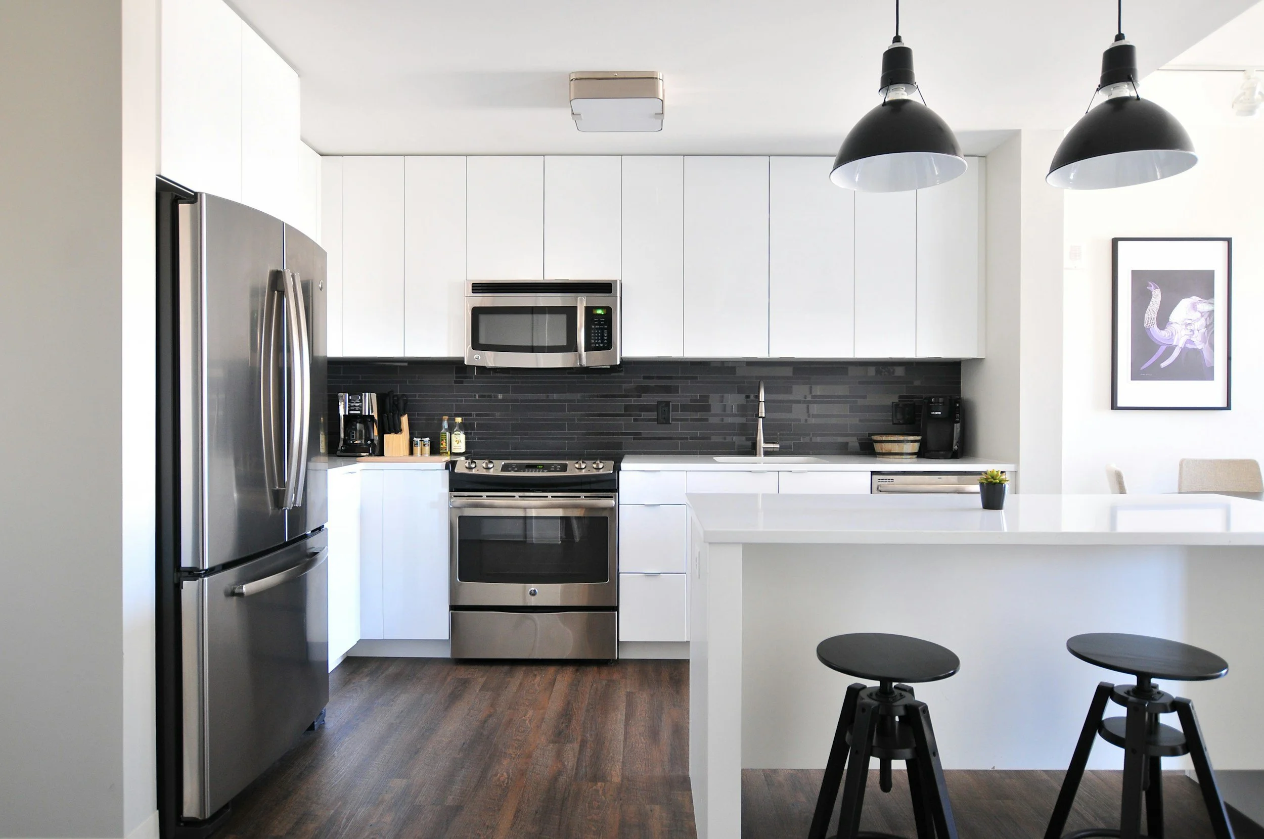 Modern kitchen with white cabinets, black backsplash, stainless steel appliances, and a white countertop island. Two black pendant lights hang from the ceiling. There are black stools at the island and a framed artwork on the wall.