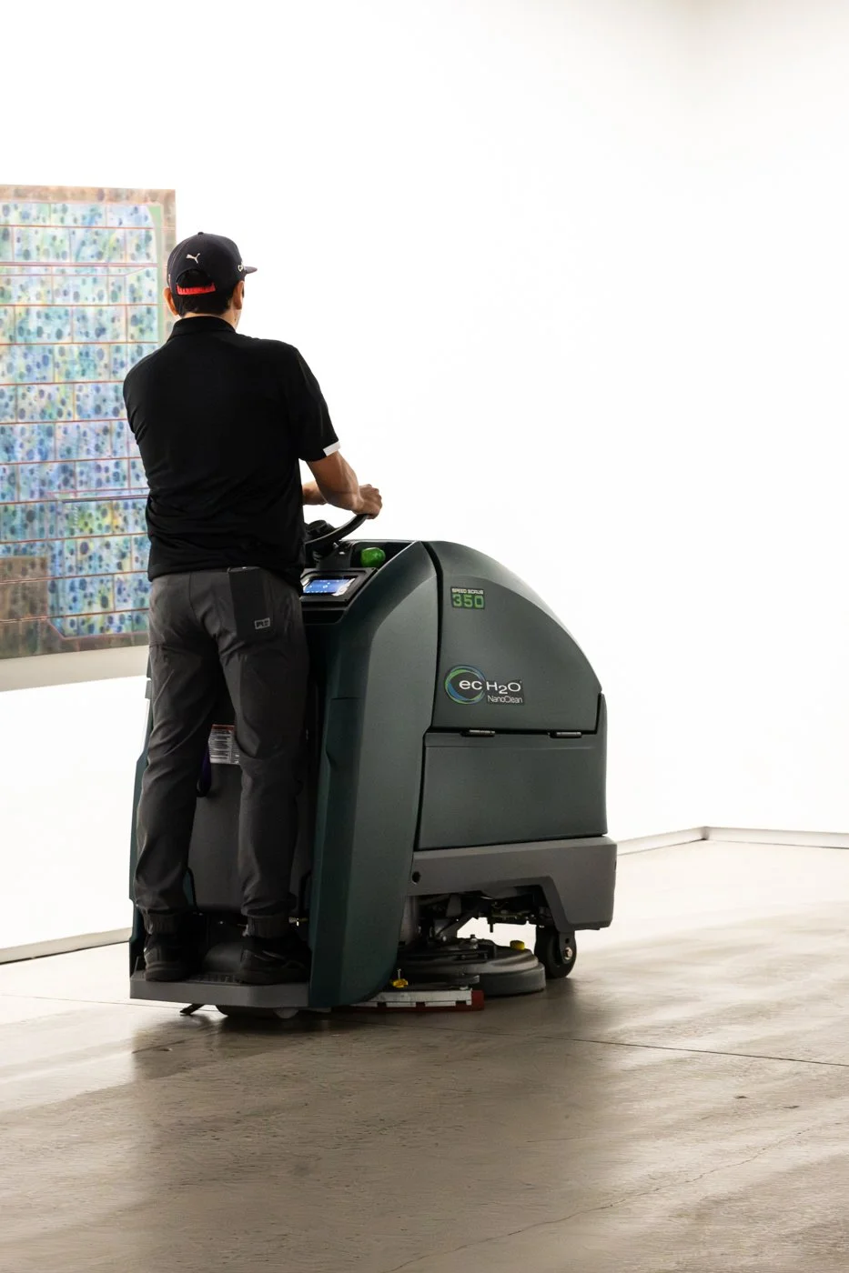 Man operating a floor cleaning machine in an art gallery, with colorful artwork hanging on the wall.