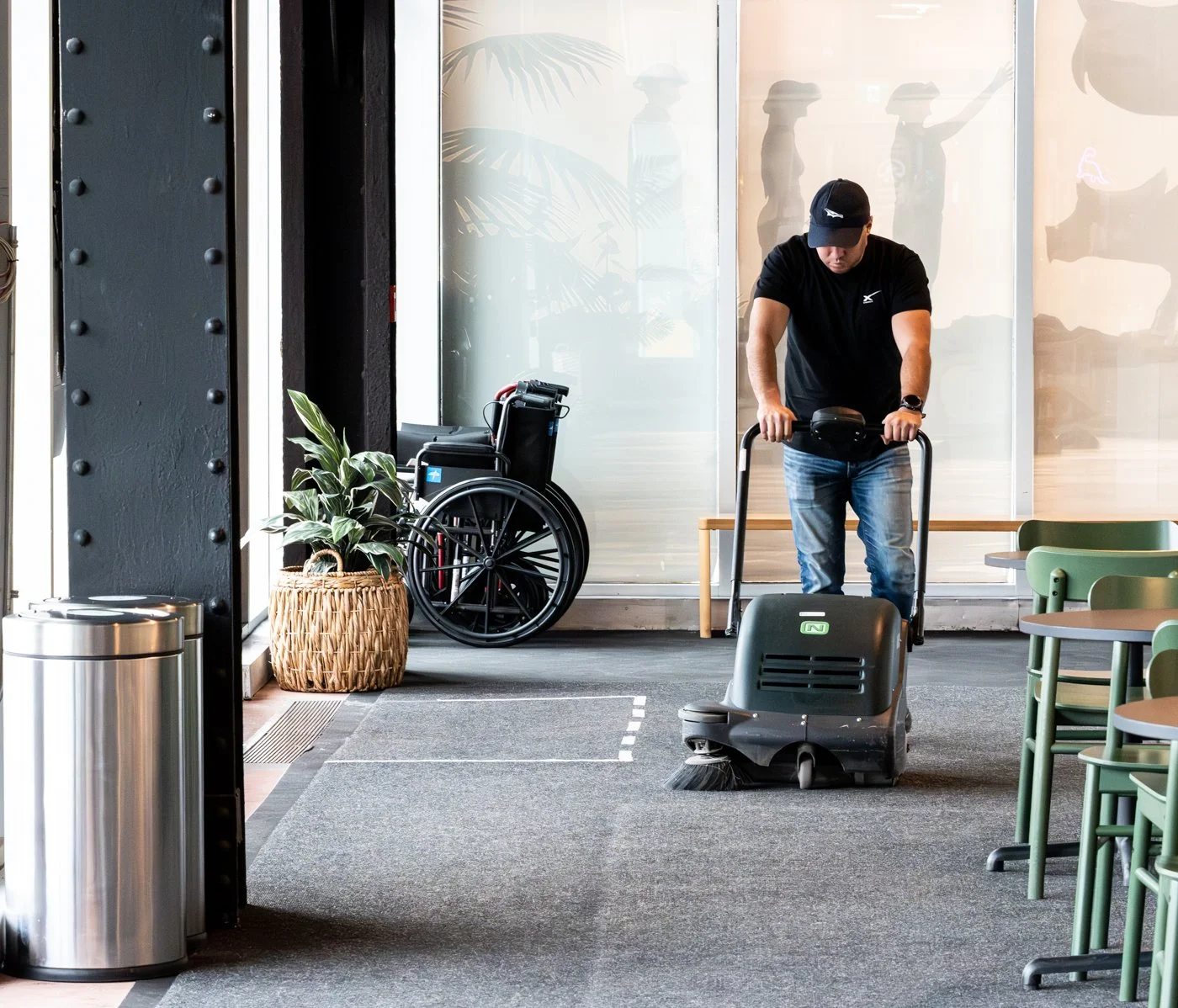 A man in a black shirt and jeans operating a floor cleaning machine in an indoor space with chairs, a wheelchair, potted plant, and glass doors