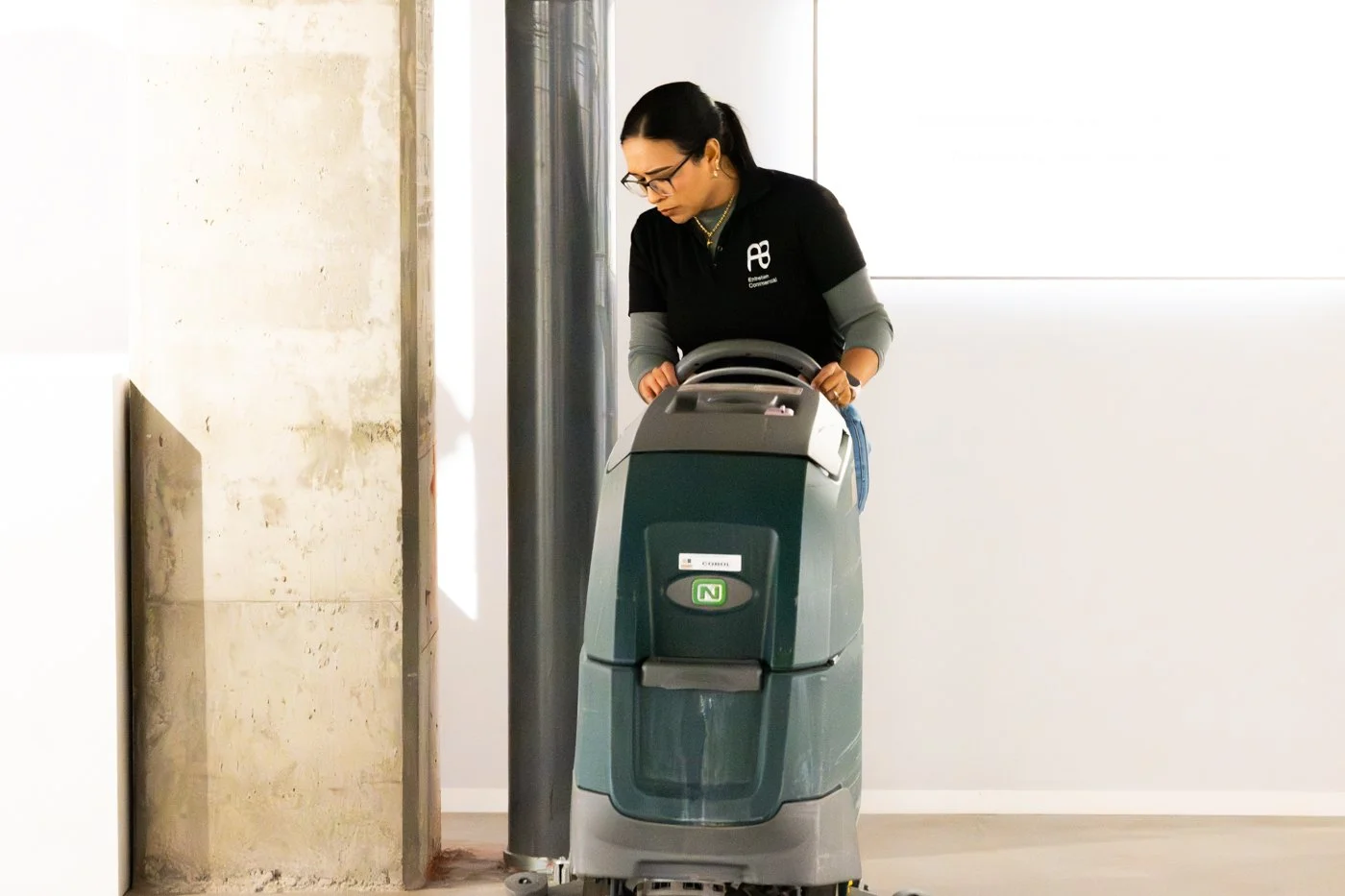 A woman wearing glasses and dark clothing operates a large commercial floor-cleaning machine in a modern indoor space.