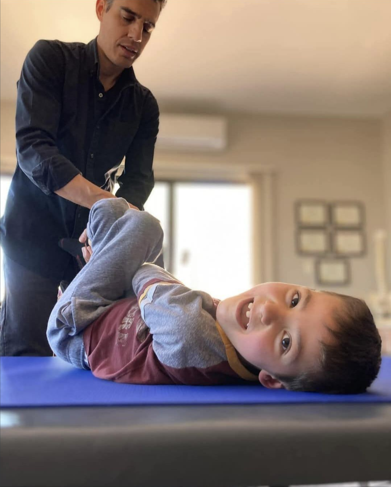 A young boy lying on his side on a blue exercise mat, smiling, as an adult assists him with stretching or physical therapy in a living room with sunlight coming through windows.