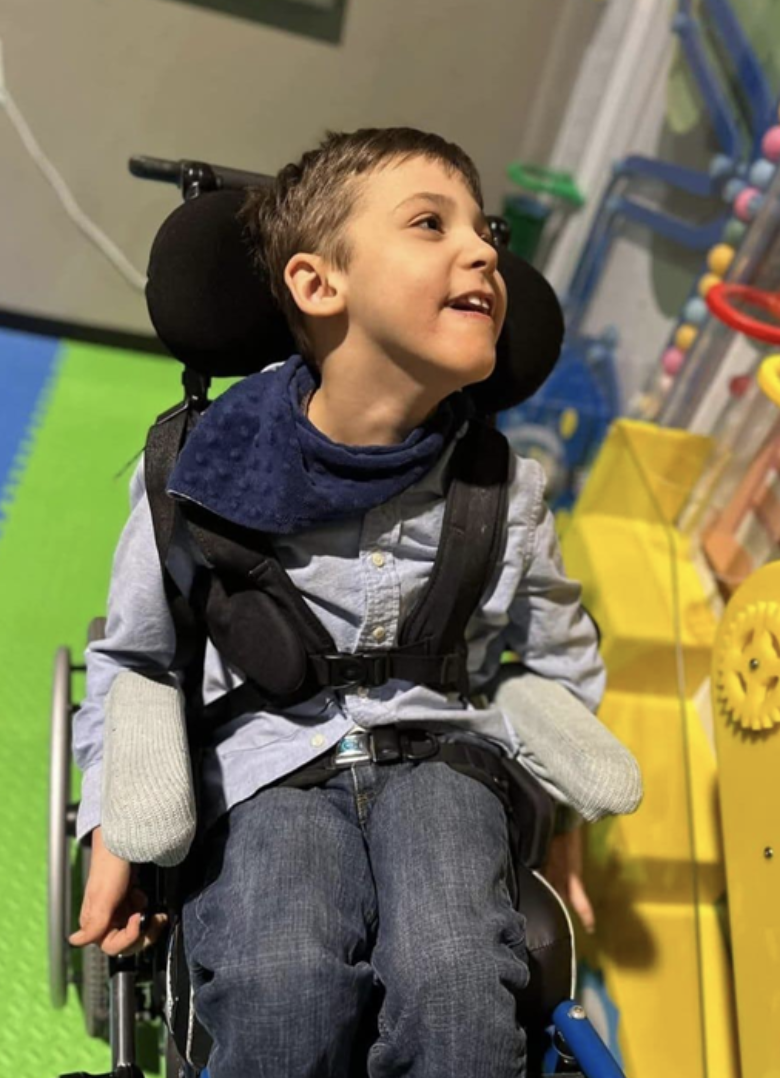 A young boy sitting in a wheelchair with a big smile, wearing a grey shirt and jeans, in a therapy or playroom setting with colorful equipment around him.