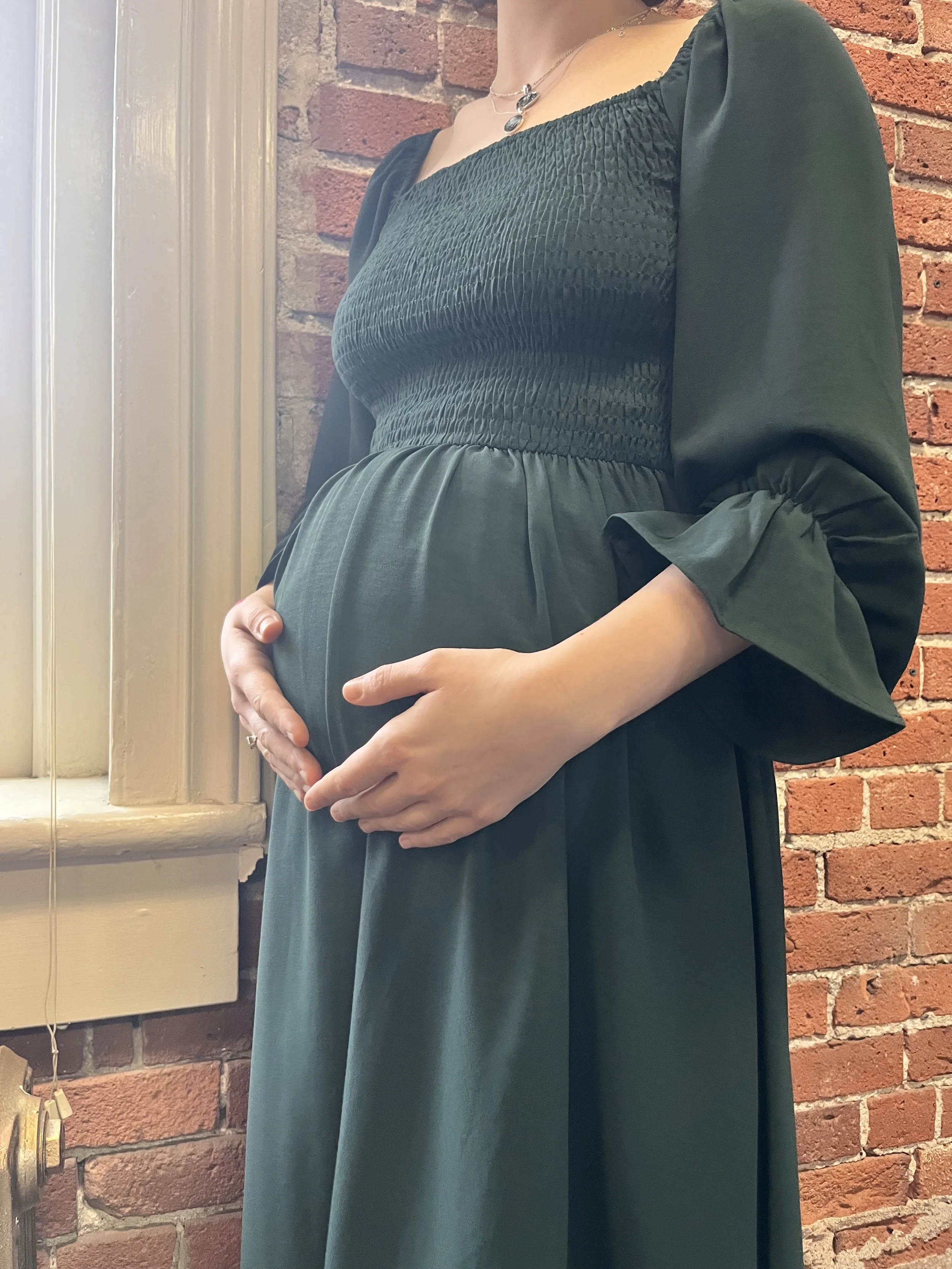Pregnant woman cradling her baby bump while standing indoors near a brick wall and a window.